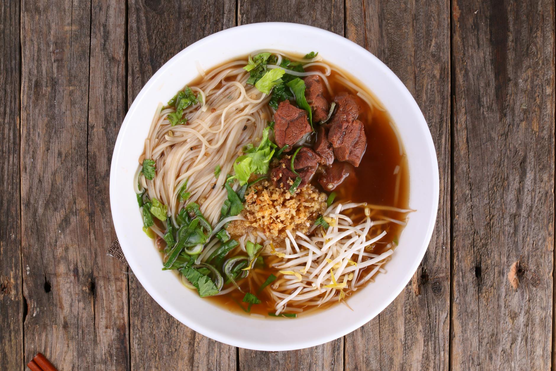 Top-down view of a deep bowl of beef noodle soup with greens, beef chunks, and chopsticks