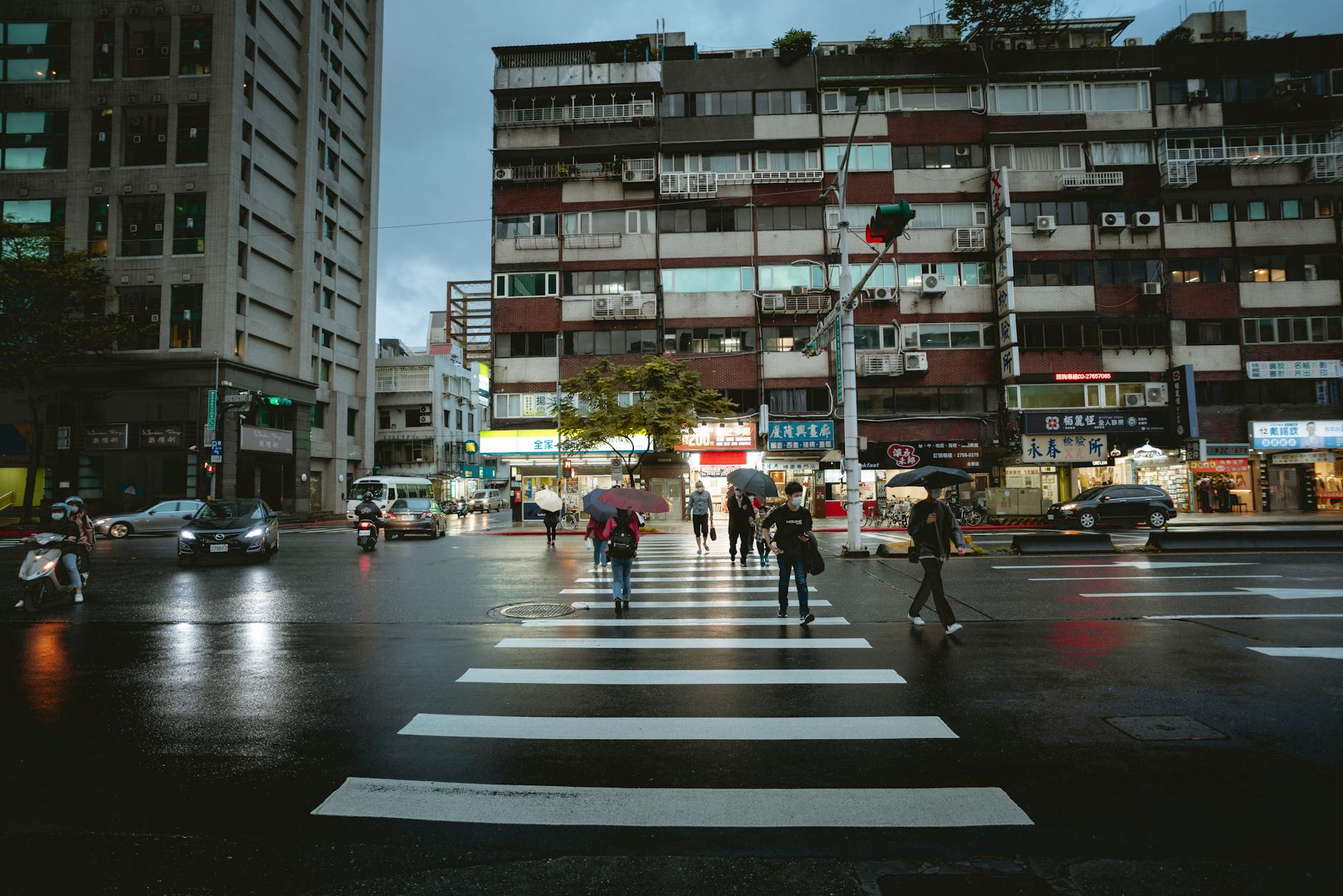 People crossing a wet city street at dusk with umbrellas and traffic