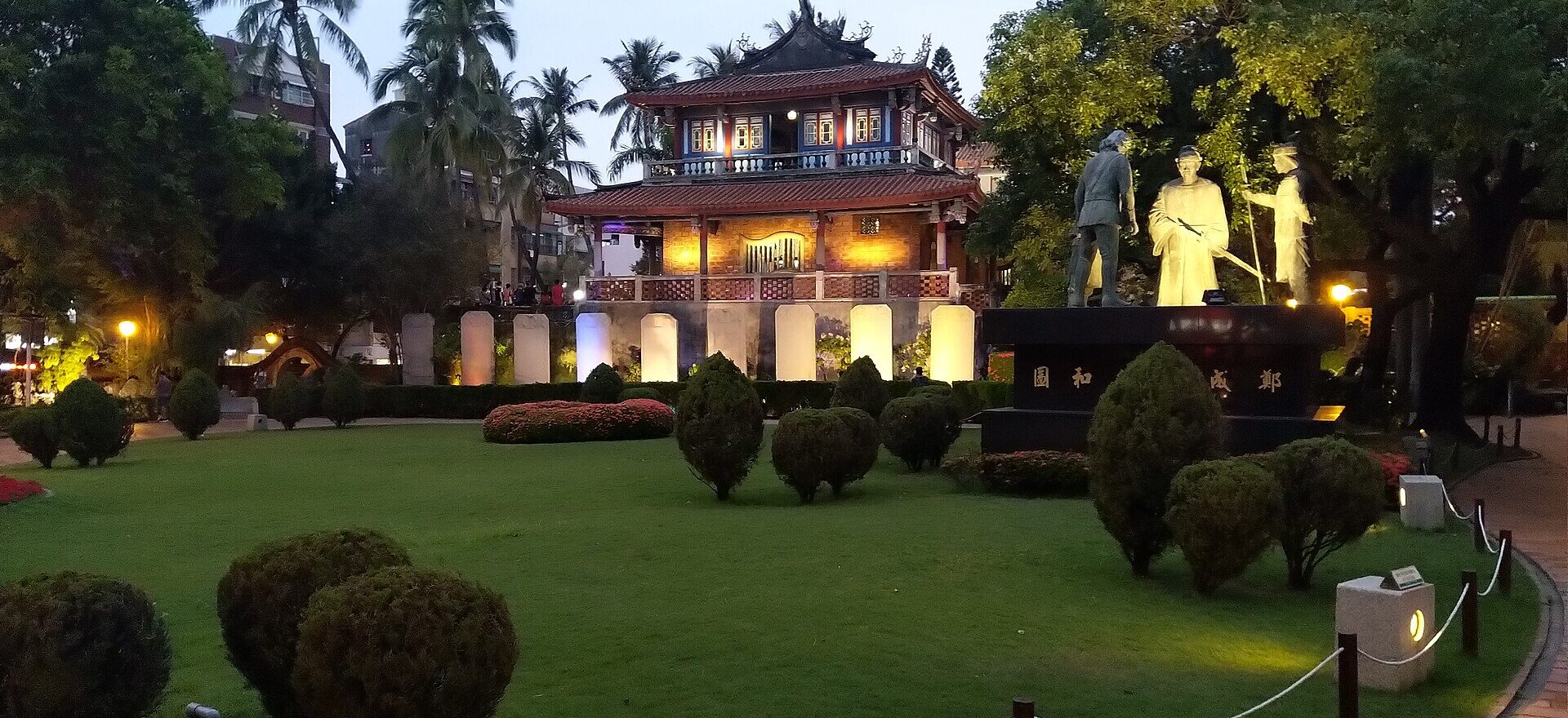 Chihkan Tower in Tainan lit at dusk with palm trees and a statue group in the garden
