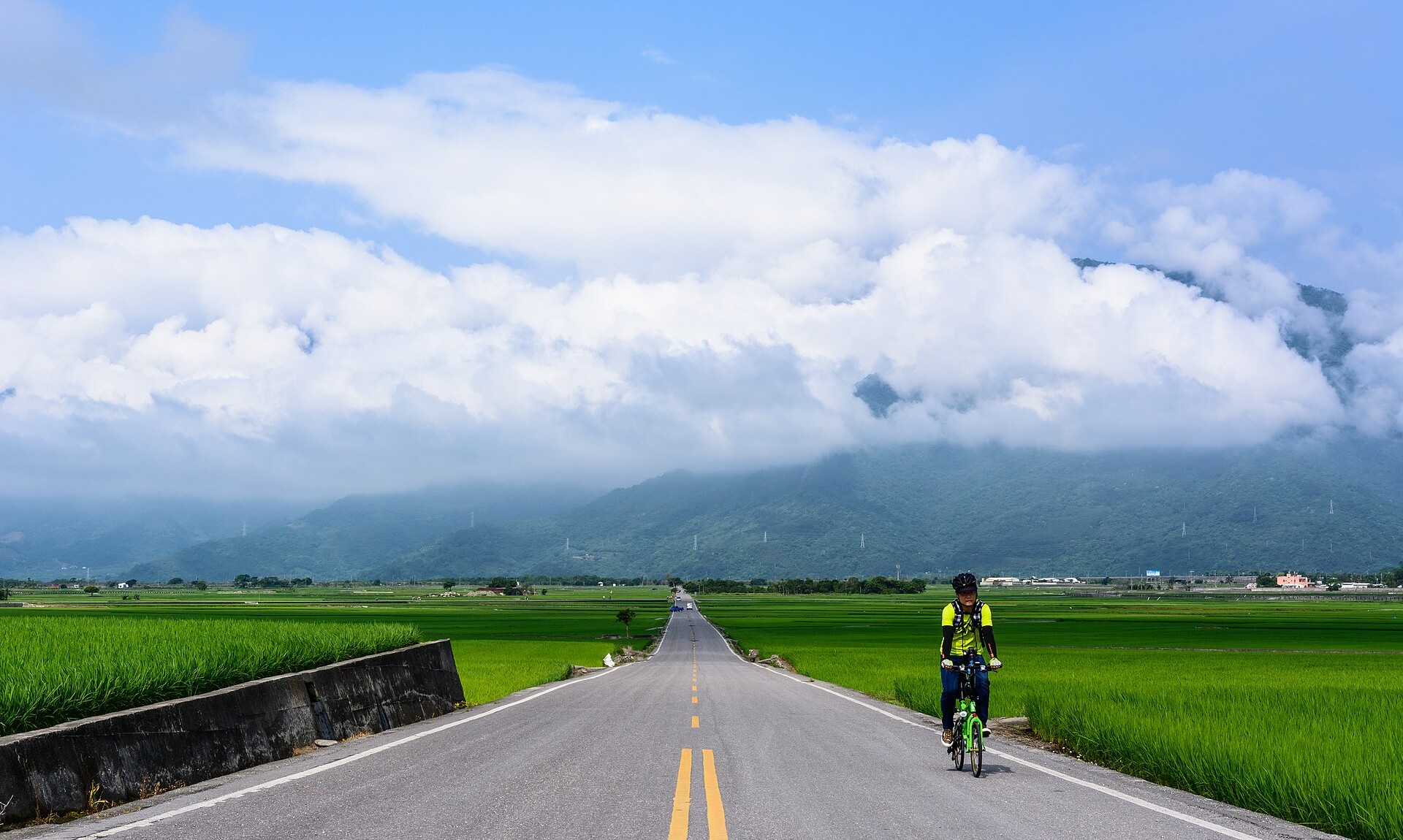 A cyclist riding along a straight road between bright green rice paddies with cloud-wrapped mountains behind, near Chishang