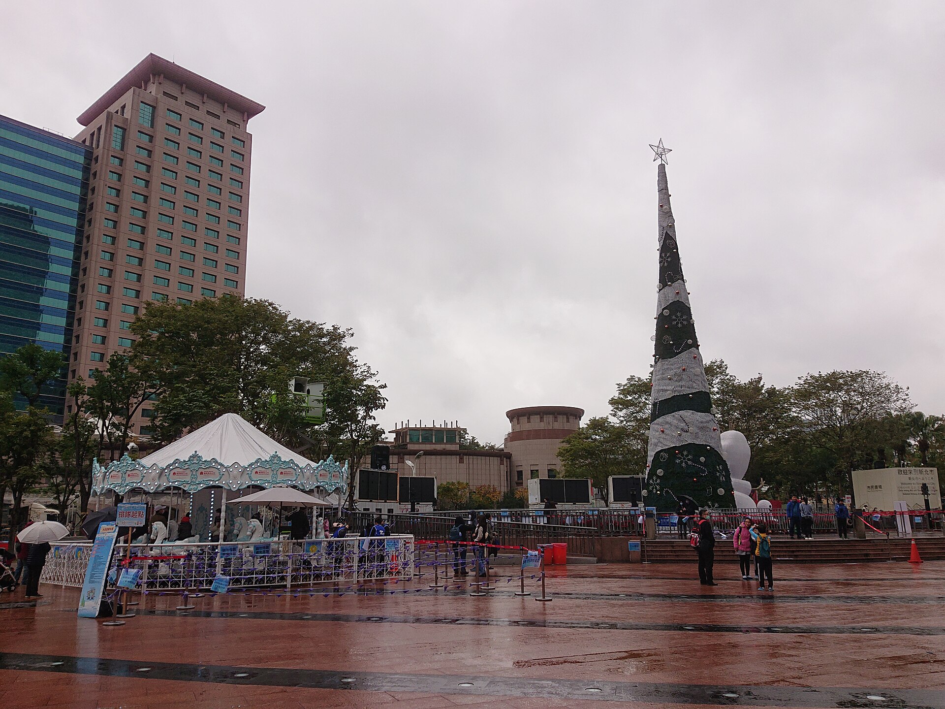 A tall Christmas tree and a carousel on the paved plaza in front of New Taipei City government buildings in Banqiao on a grey, wet day