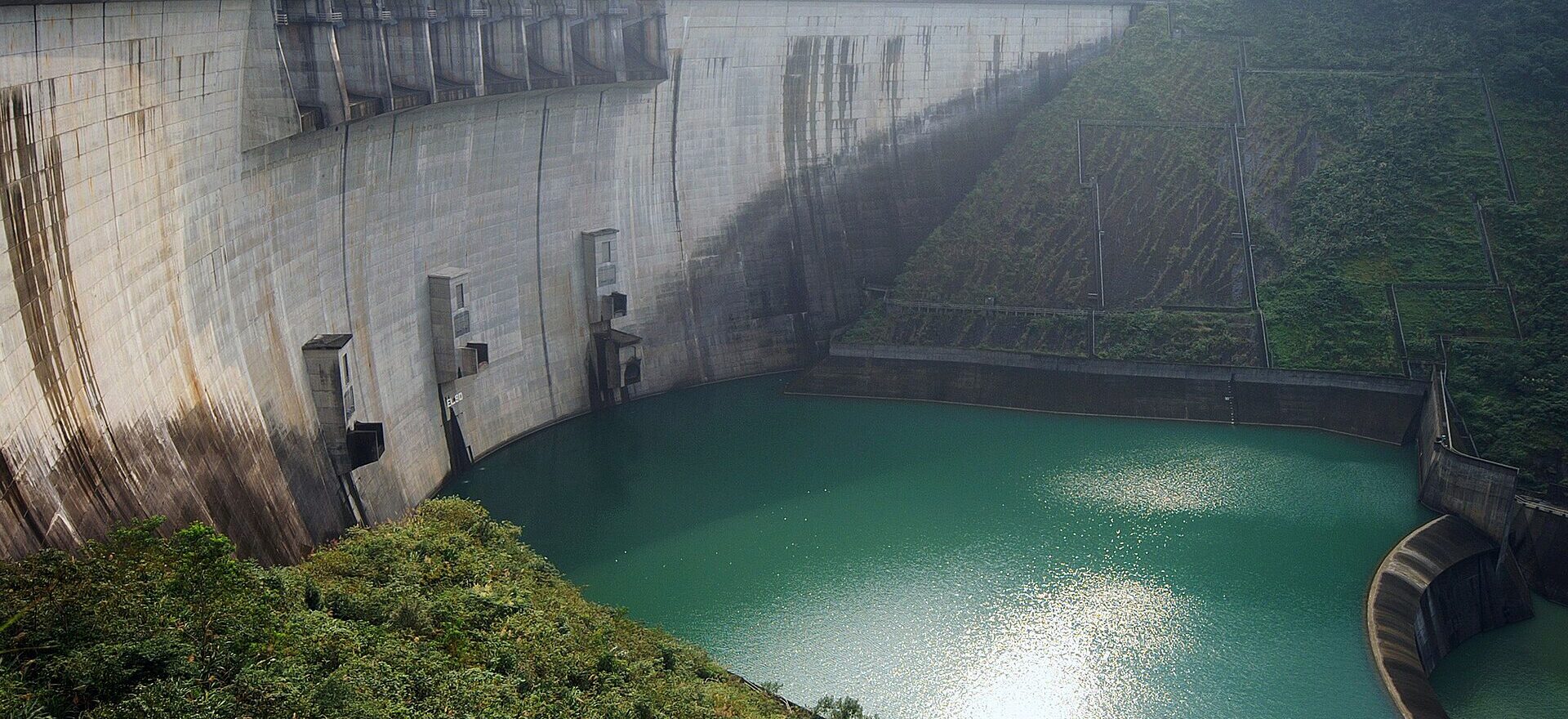 The curved concrete dam wall of Feitsui Reservoir with green water and forested hills