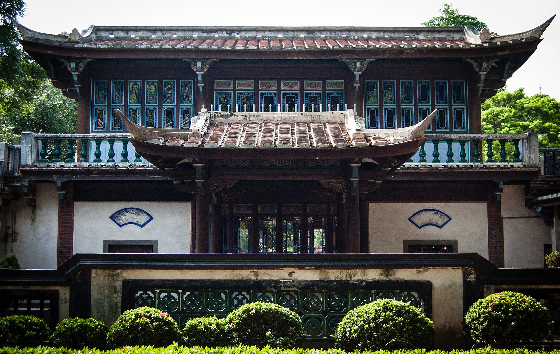 The two-storey Guanjia Tower in the Lin Family Garden, with an upturned tiled roof, blue lattice windows, balustrades and fan-shaped wall panels behind clipped shrubs