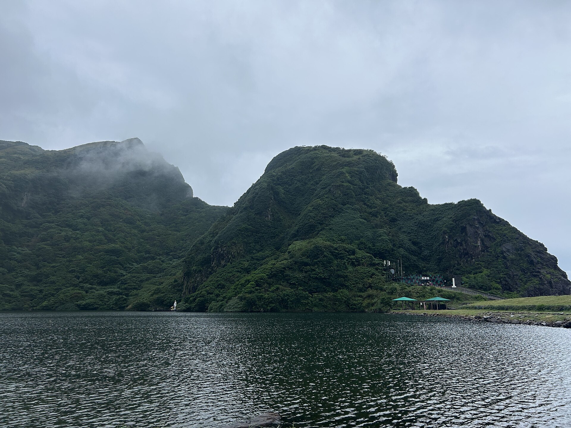 A dark lake below steep green volcanic hills on Guishan Island under low grey cloud, with pavilions on the shore
