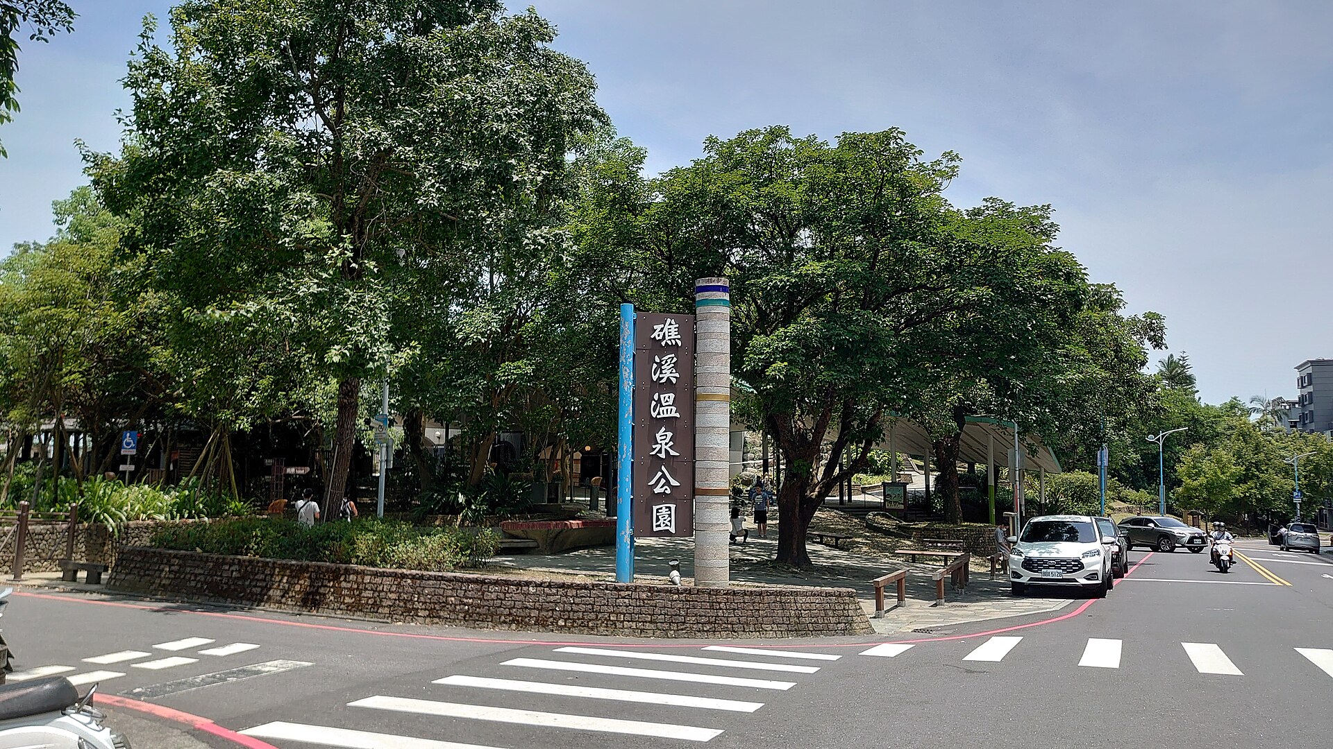 The entrance to Jiaoxi Hot Spring Park with its tall brown sign in Chinese characters, shade trees and a quiet street crossing