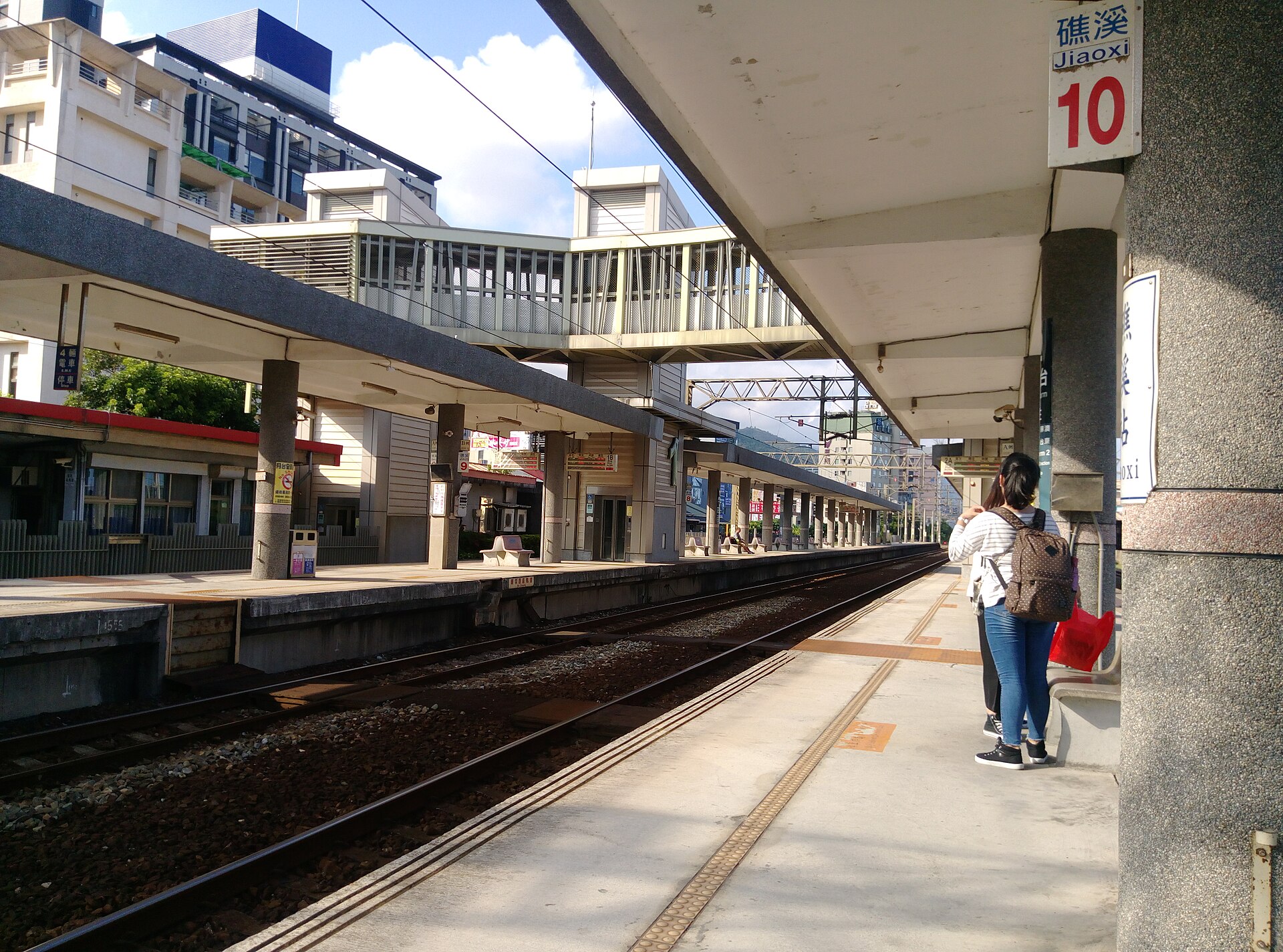 The platform at Jiaoxi station with a blue and white station sign, a covered footbridge and hotel blocks beyond the tracks