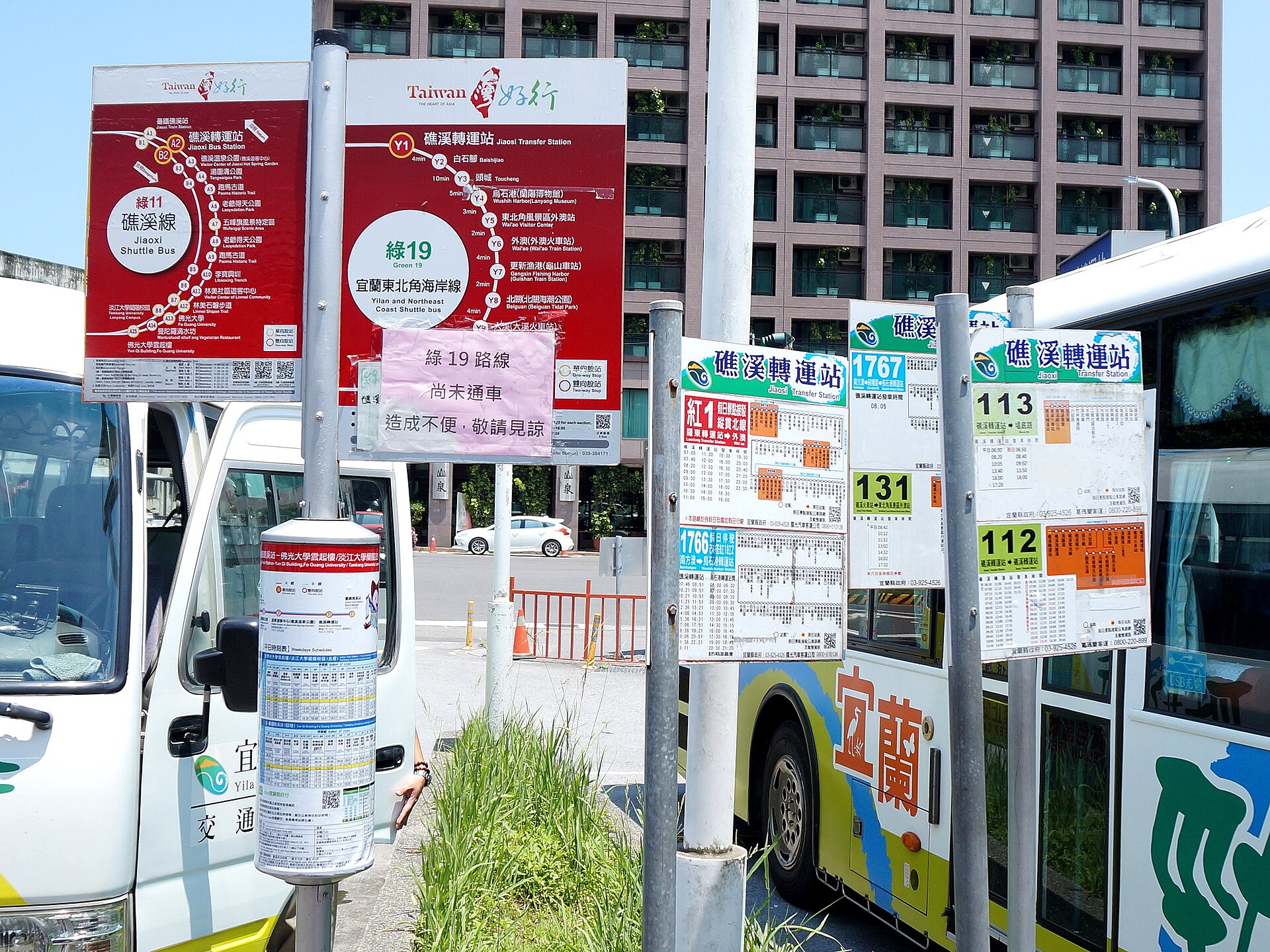 Tourist shuttle route maps and local bus timetables on poles at Jiaoxi Transfer Station, with a Yilan bus parked alongside