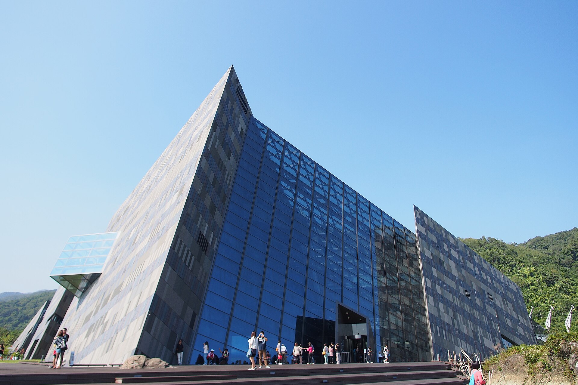 The angular glass and stone wedge of the Lanyang Museum rising against a clear blue sky with visitors at the entrance
