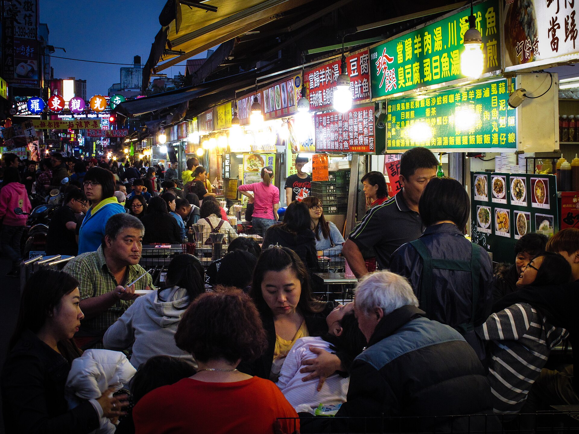 Families eating at crowded tables beneath brightly lit stall signs at Luodong Night Market at dusk