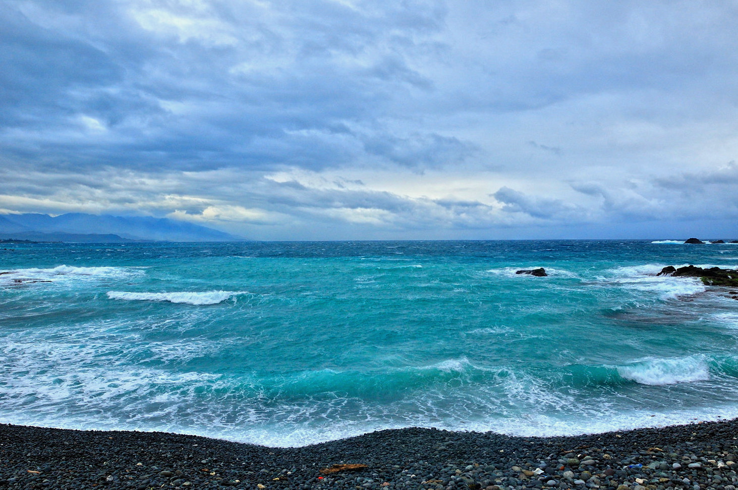 Turquoise waves breaking on a dark pebble beach under a heavy grey sky at Sanxiantai on the east coast