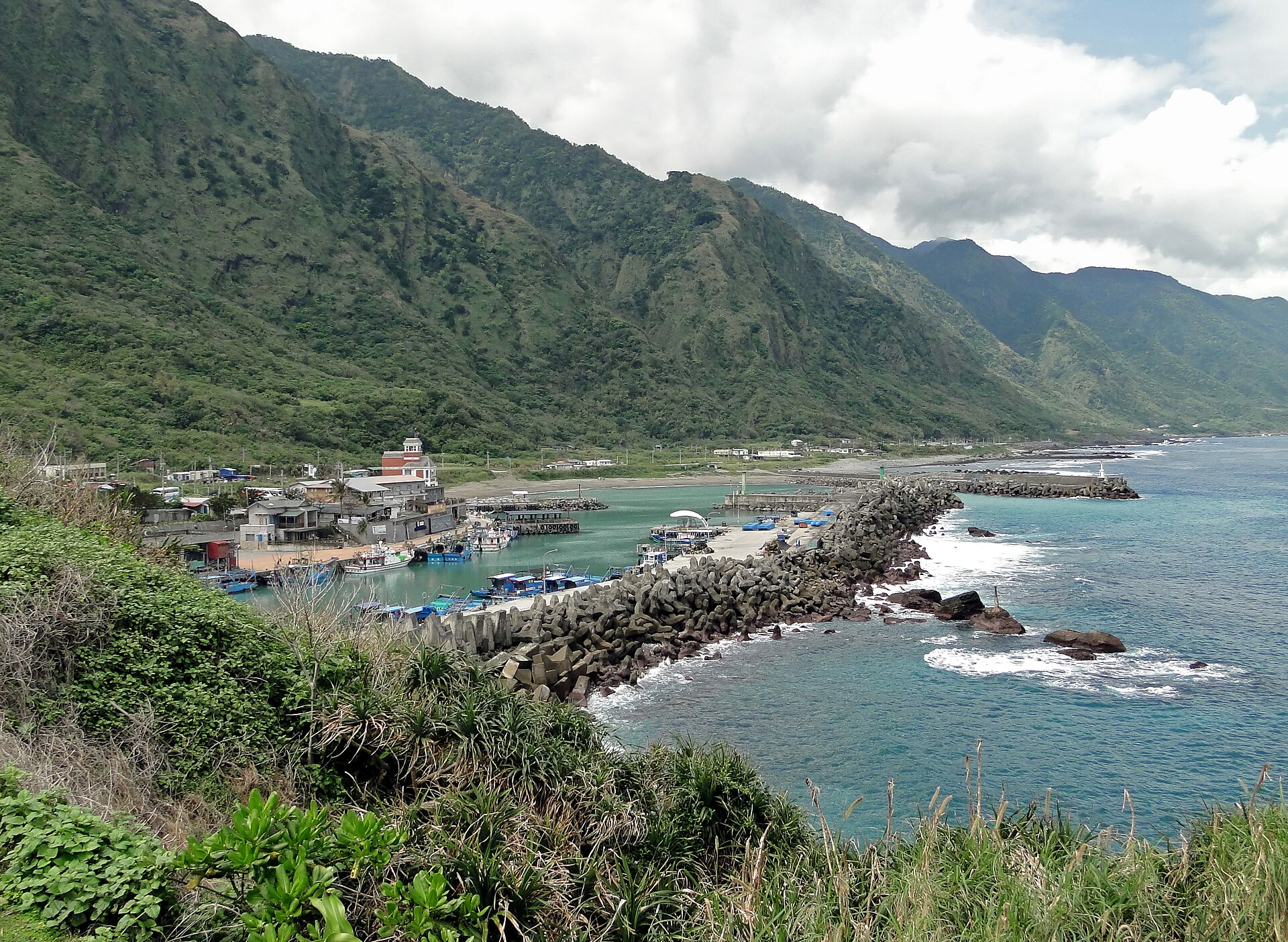 Shitiping fishing harbour below steep green mountains, with boats inside a breakwater and waves breaking on dark rocks