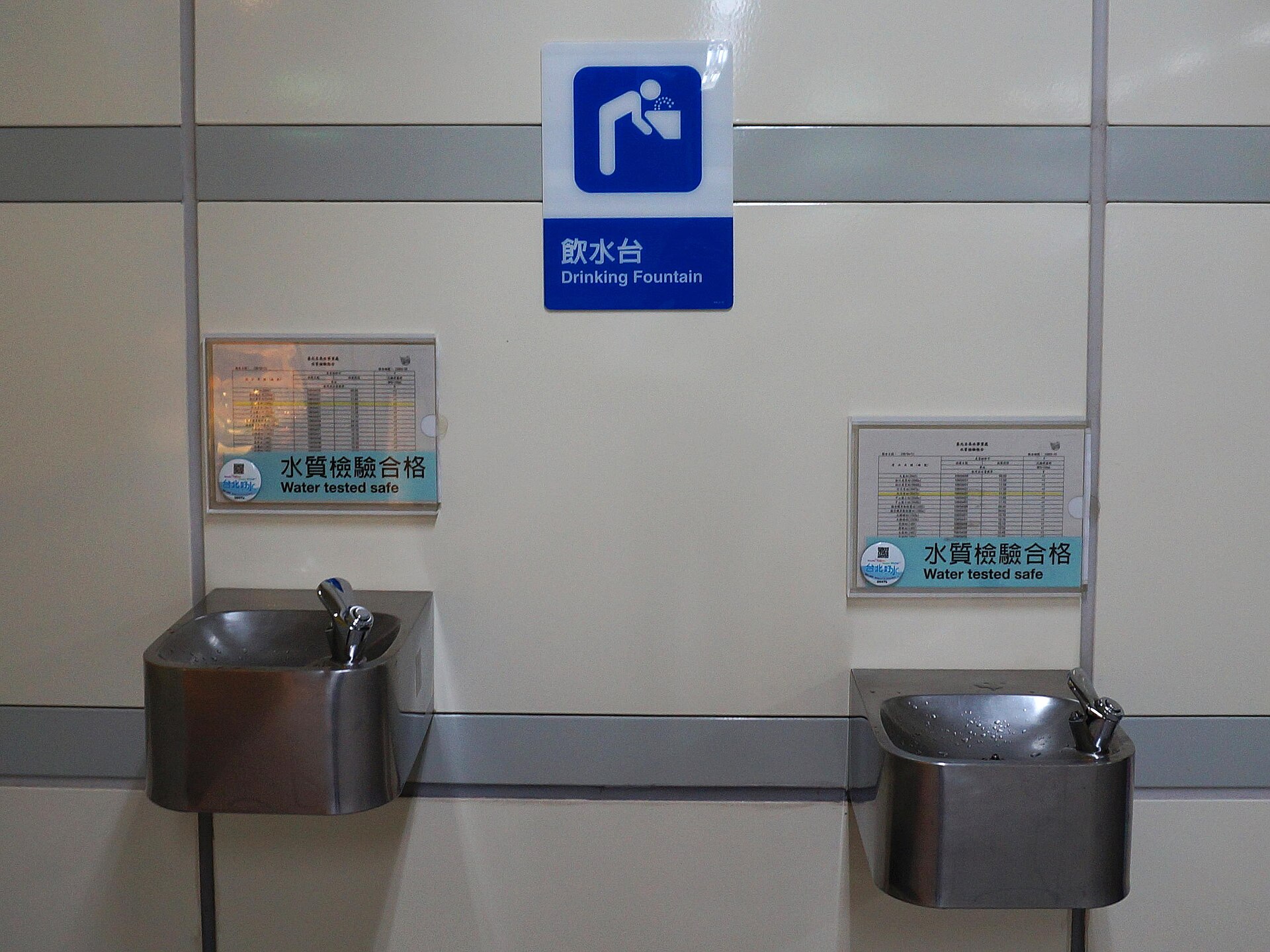 Two stainless steel drinking fountains in a Taipei MRT station beneath a bilingual sign, each with a laminated water quality test certificate