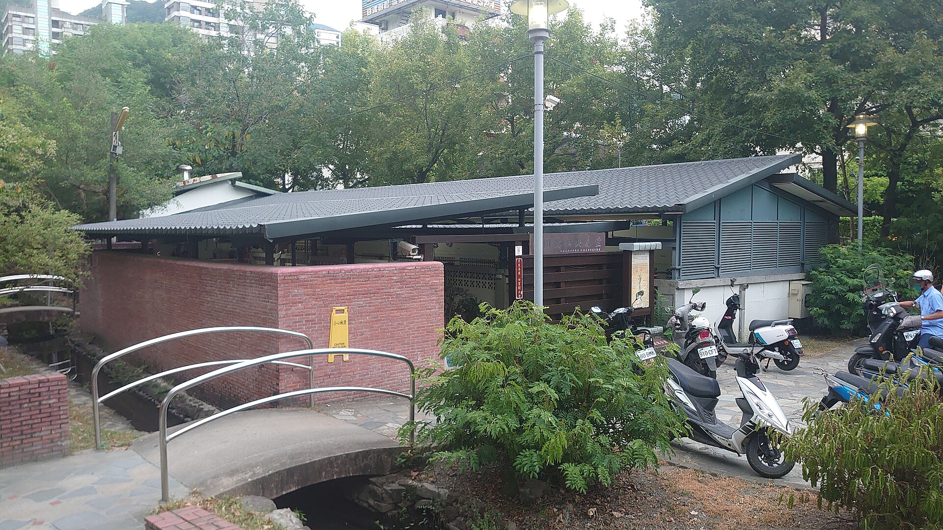 The low-roofed public bathhouse at Tangweigou with a red brick wall, a small arched bridge over the hot spring stream and scooters parked outside