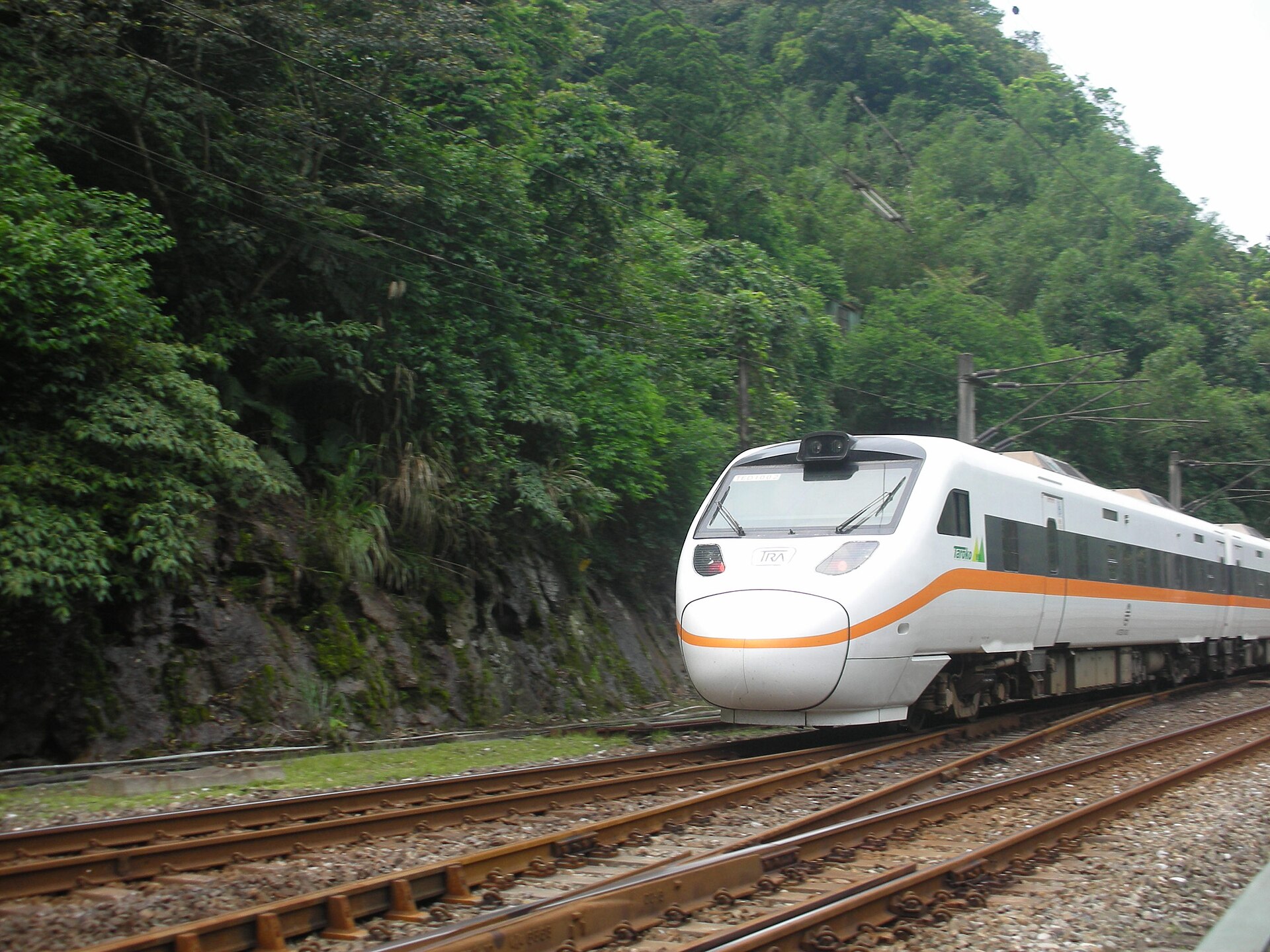 A white Taroko Express tilting train with an orange stripe running past a steep forested hillside