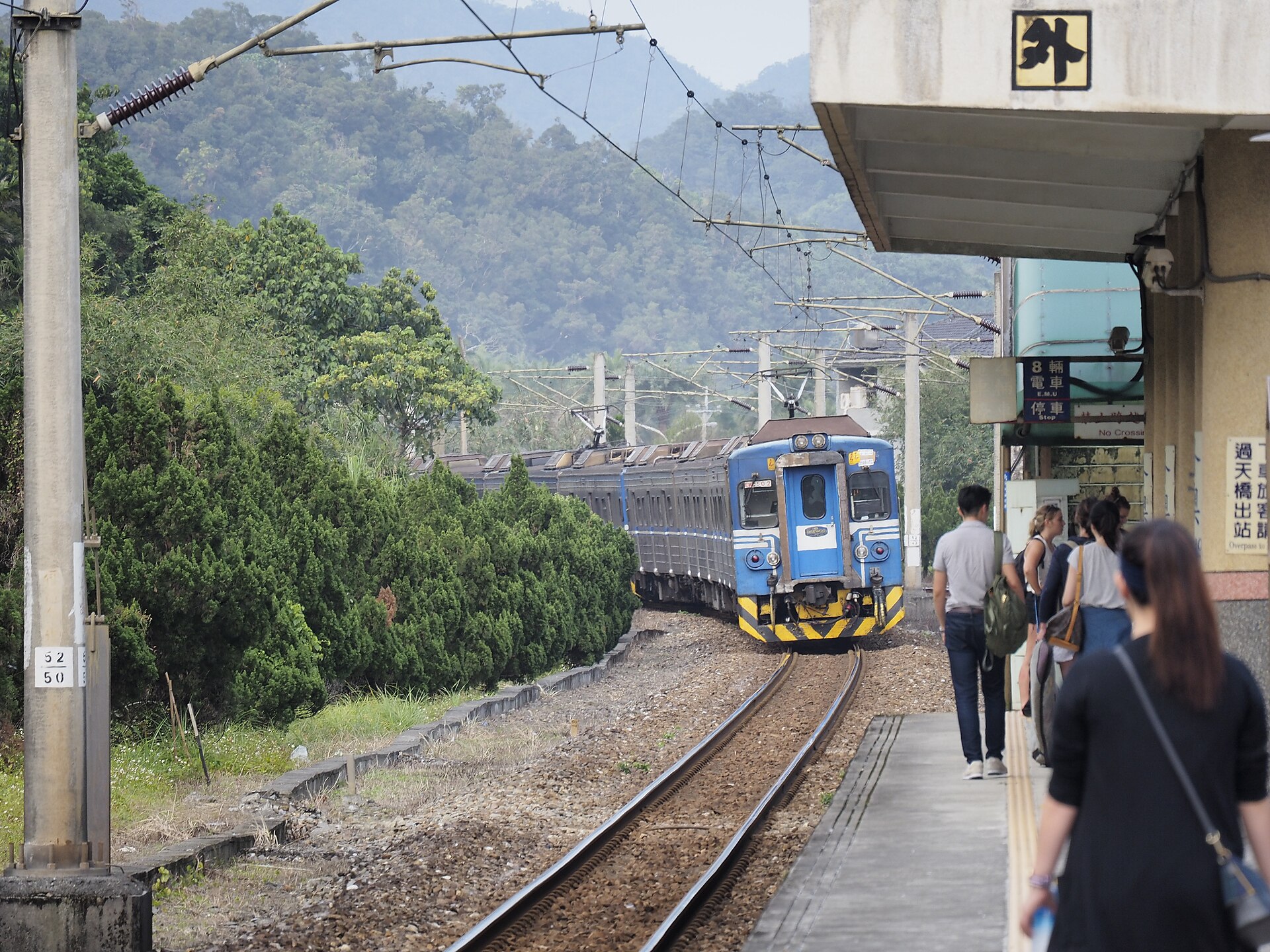 A blue local train rounding the curve into Wai'ao station with passengers waiting on the platform and wooded hills behind