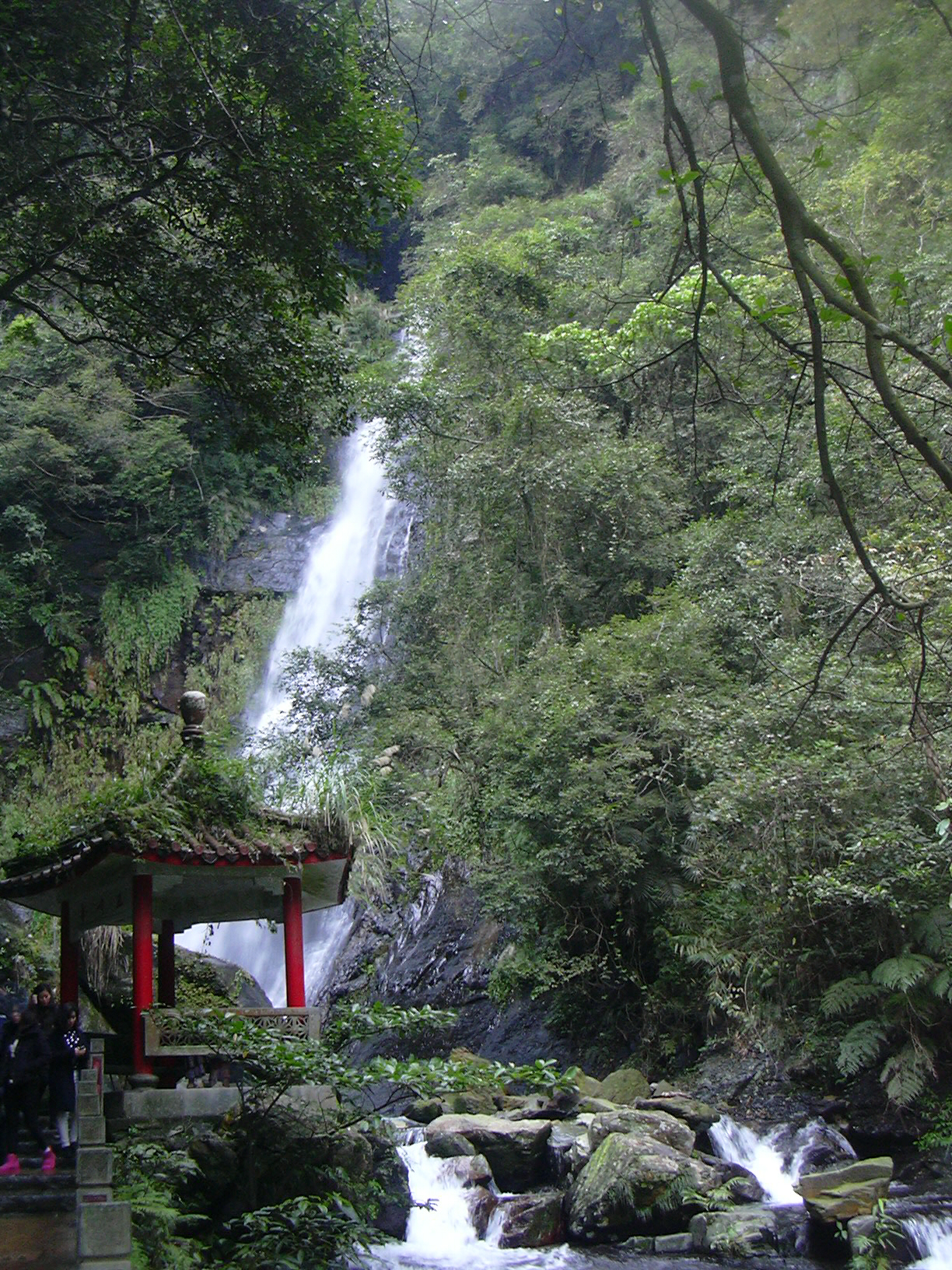 A tall white waterfall dropping through dense green forest behind a red-pillared pavilion and a rocky stream