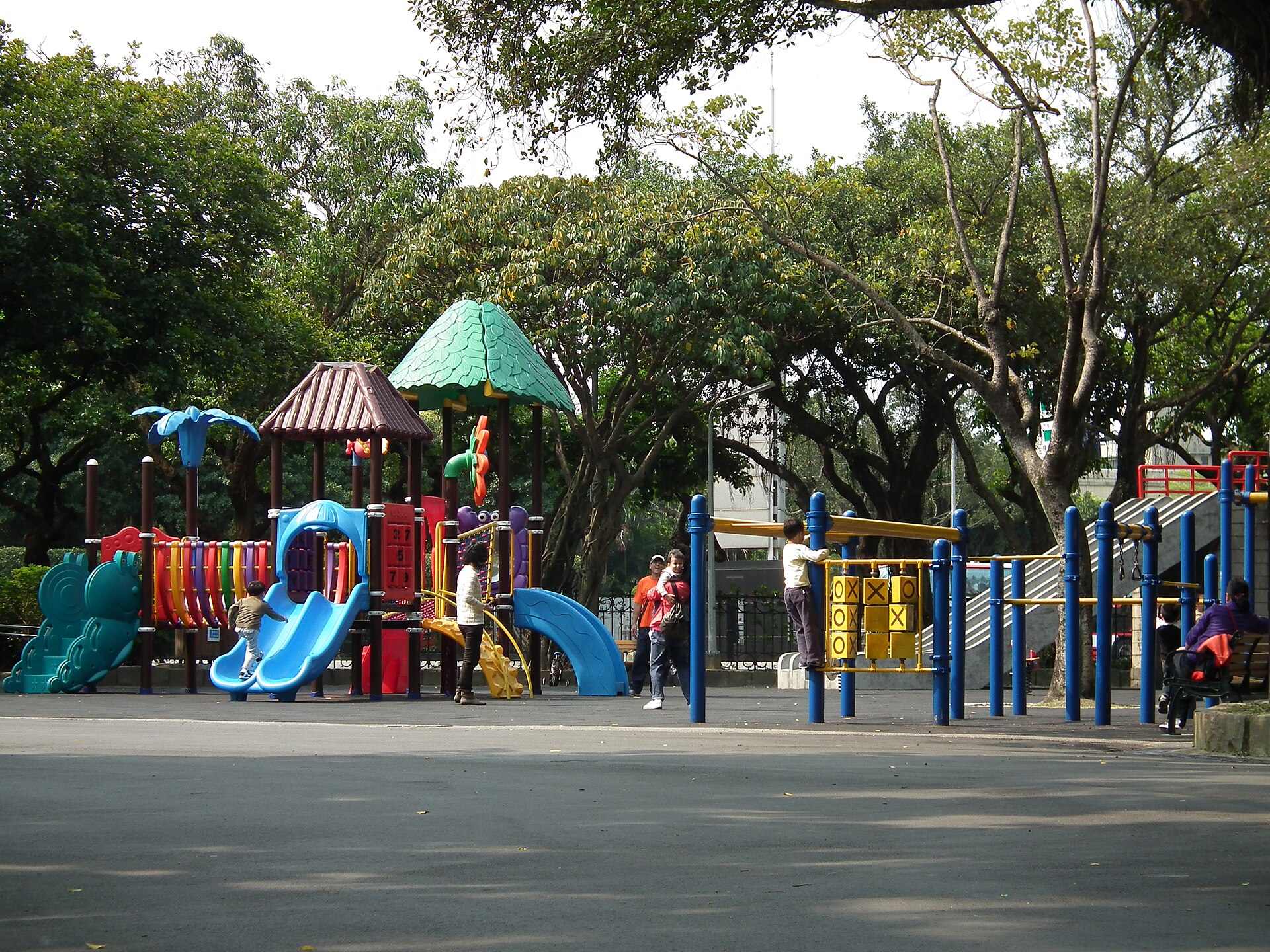 Adults and children use colorful slides and climbing equipment beneath trees at Taipei's 228 Peace Memorial Park.