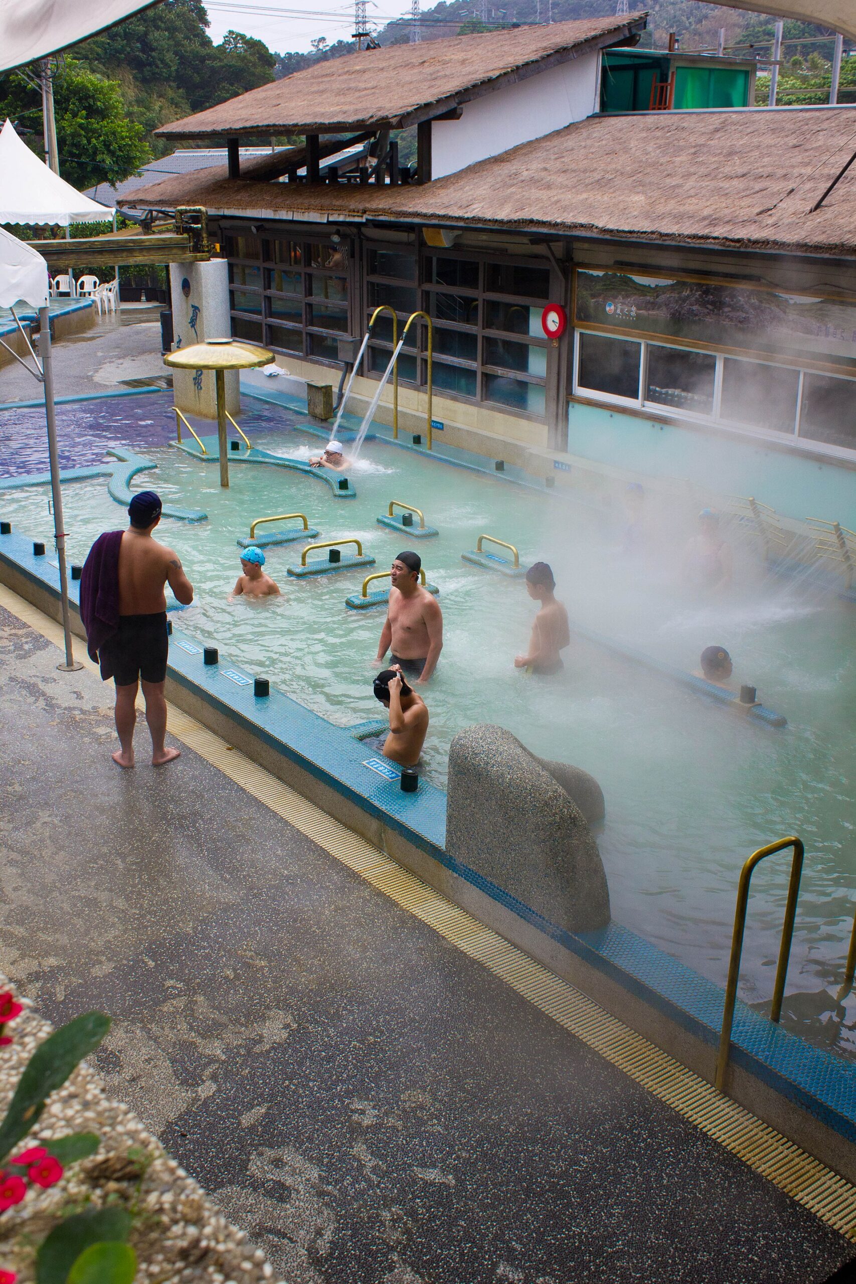 Adults wearing swimwear and swimming caps bathe in a steaming outdoor pool beside a low building in Beitou.
