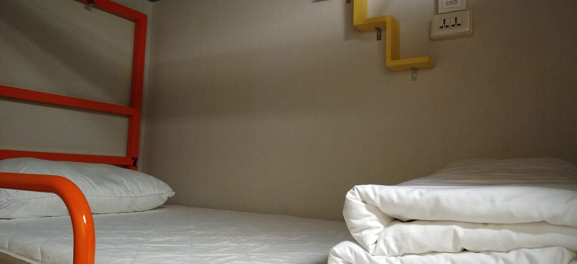 A bunk at Homey Hostel in Taipei has white bedding, an orange safety rail and a small yellow shelf.