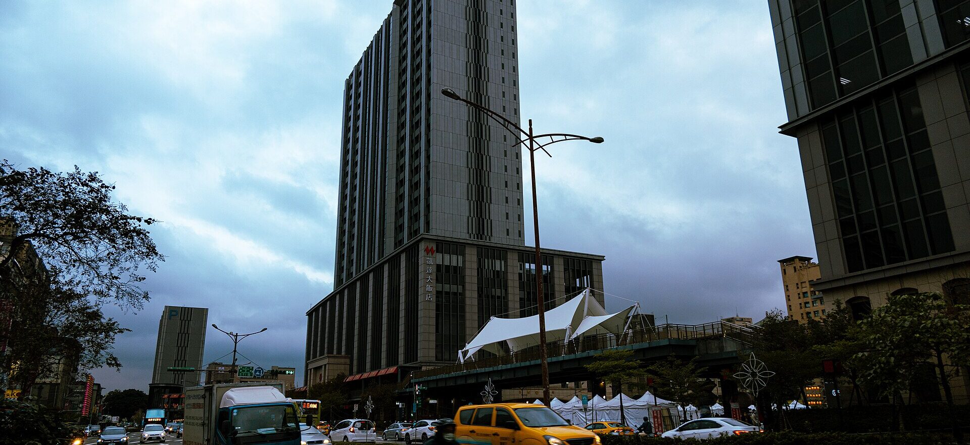 Caesar Metro Taipei towers over a busy Taipei street with taxis, scooters and a white canopy beside the building.