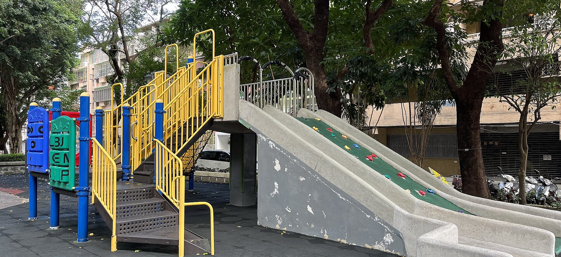 A concrete slide, climbing slope and yellow stair rails sit beneath mature trees in a Taipei neighborhood park.