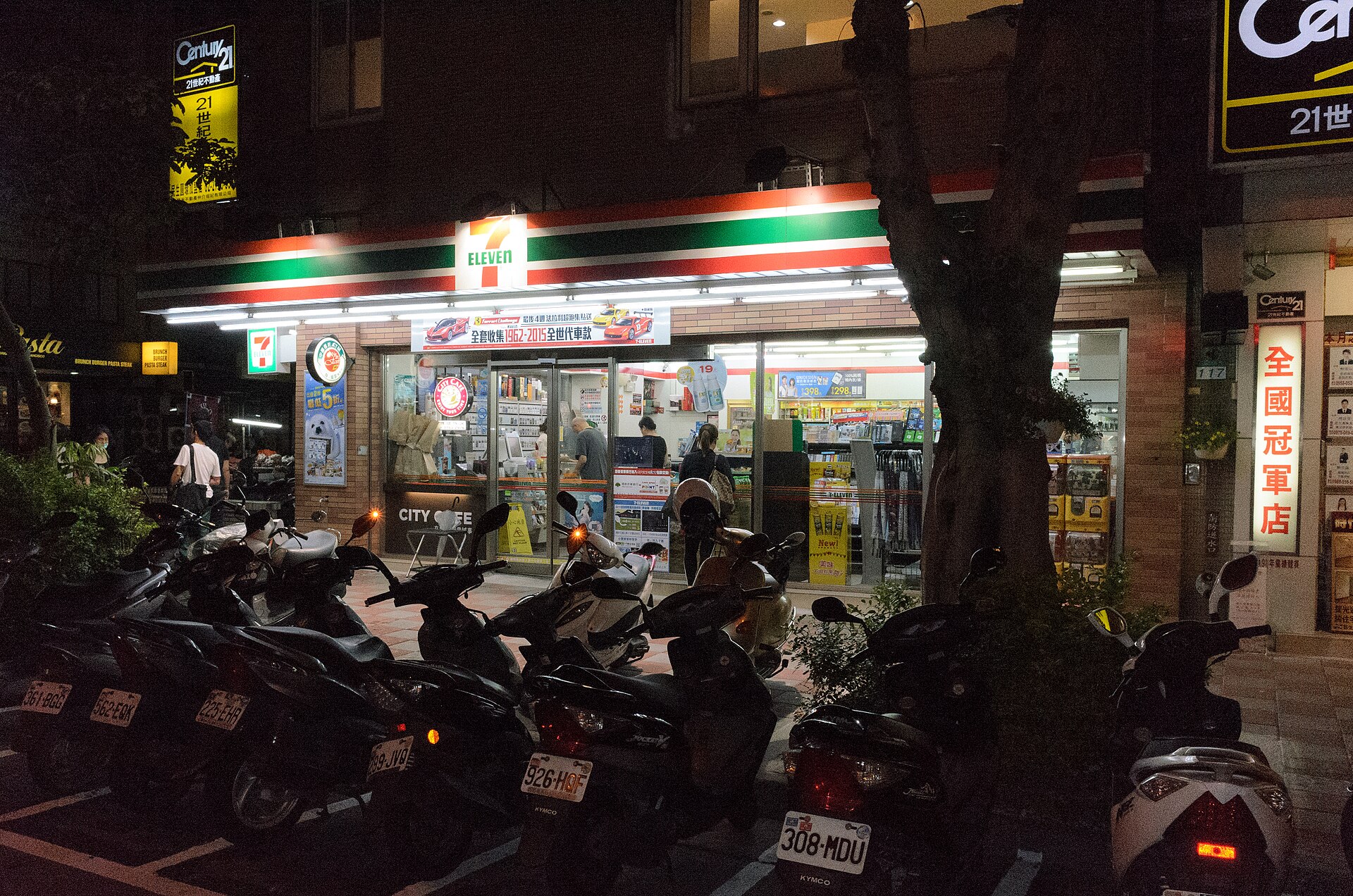 Customers stand inside a brightly lit 7-Eleven in Taipei at night, with parked scooters lining the pavement.