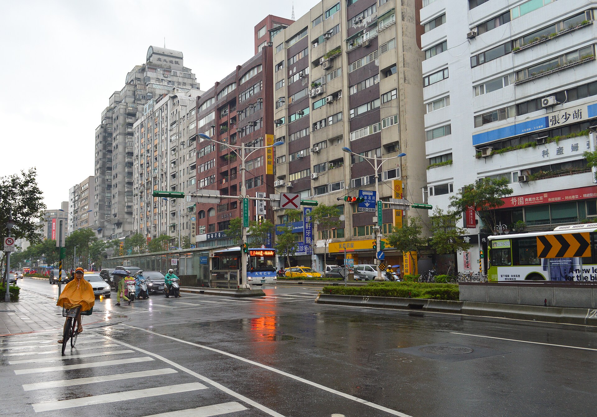 A cyclist in a yellow rain poncho approaches a wet crossing on Taipei's Xinyi Road, alongside scooters, cars and buses.