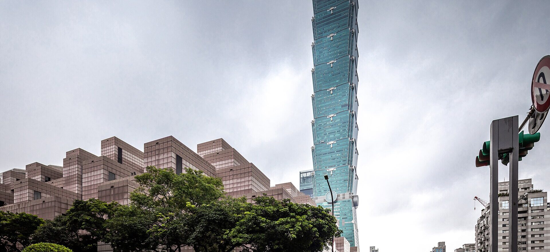 Dark rain clouds hang above Taipei 101 and the World Trade Center, while pedestrians and scooter riders cross wet Xinyi Road.