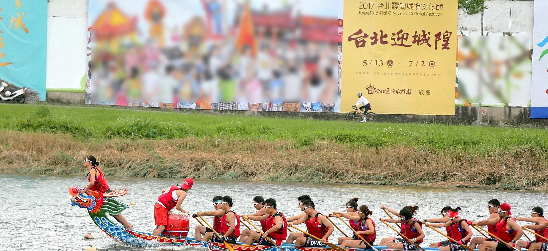 A dragon boat crew in red life jackets paddles along the Keelung River in Taipei, behind a brightly painted dragon head.