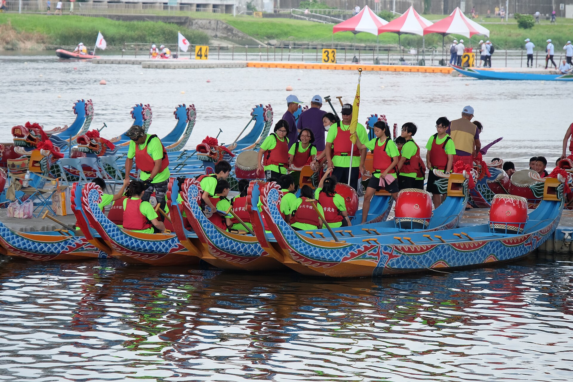 Dragon boat crews wearing bright green shirts board decorated boats with red drums at a Taipei riverside pier.