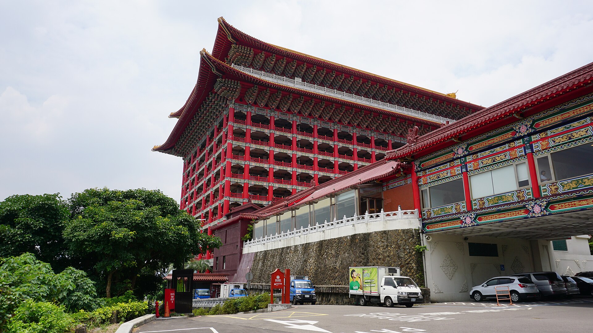 The Grand Hotel in Taipei rises behind its entrance drive, with red balconies, an ornate roof and trees beside parked vehicles.