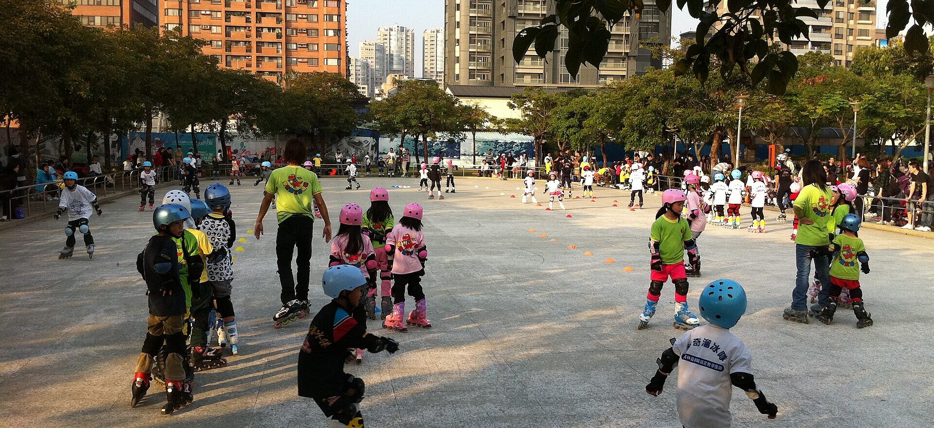 Helmeted children practice inline skating with adults on a broad paved area in Daan Forest Park, Taipei, surrounded by city buildings.