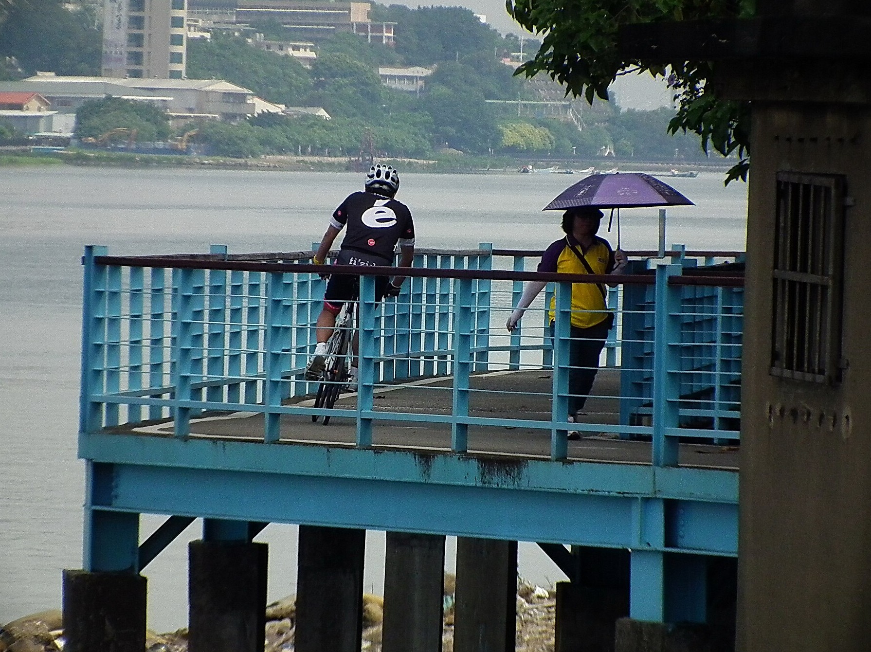 A helmeted cyclist follows the blue-railed Tamsui Golden Shore path beside the river, passing a pedestrian with a parasol.