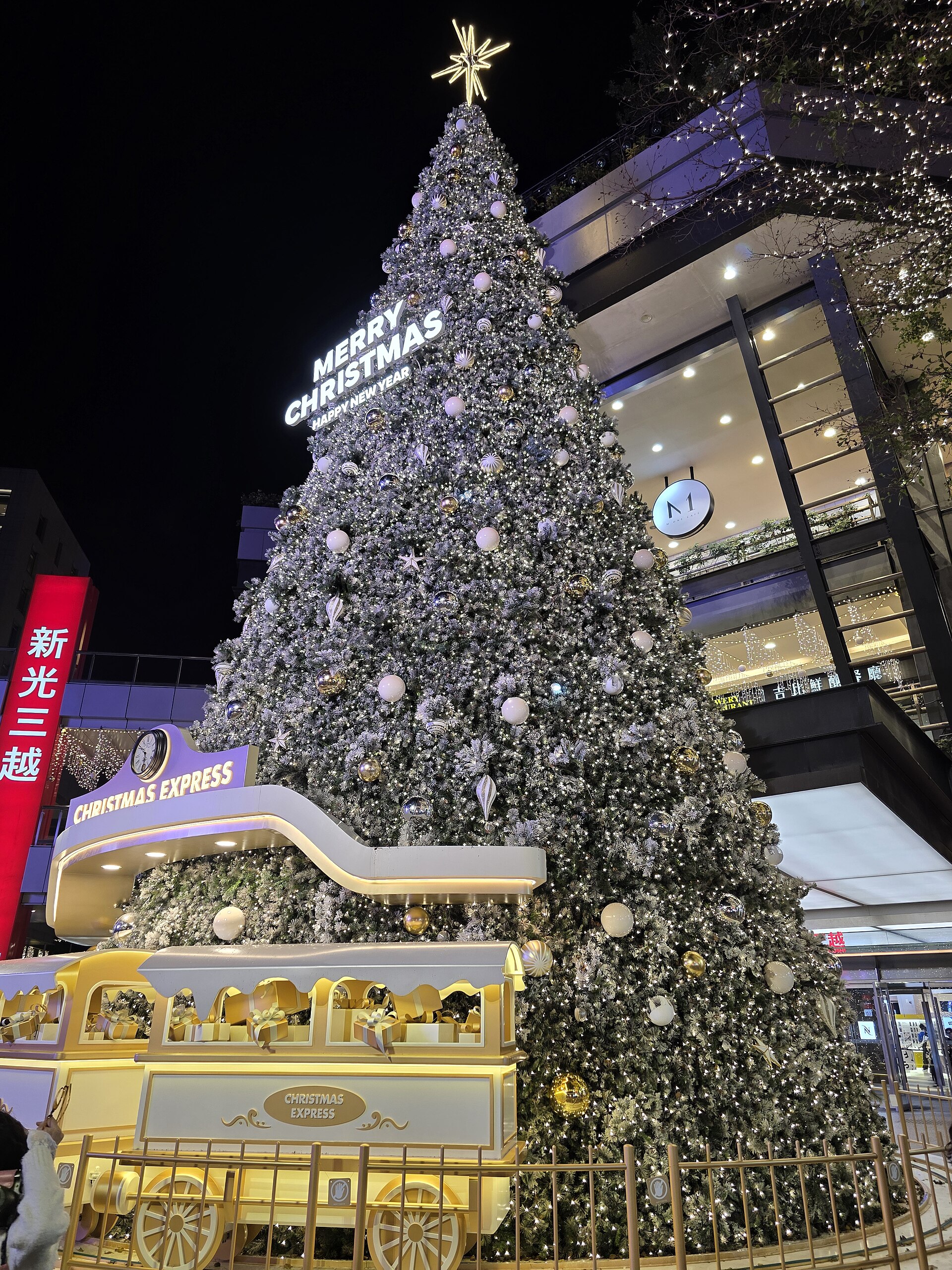 An illuminated Christmas tree and yellow decorative train stand outside Shin Kong Mitsukoshi Xinyi Place A11 in Taipei at night.