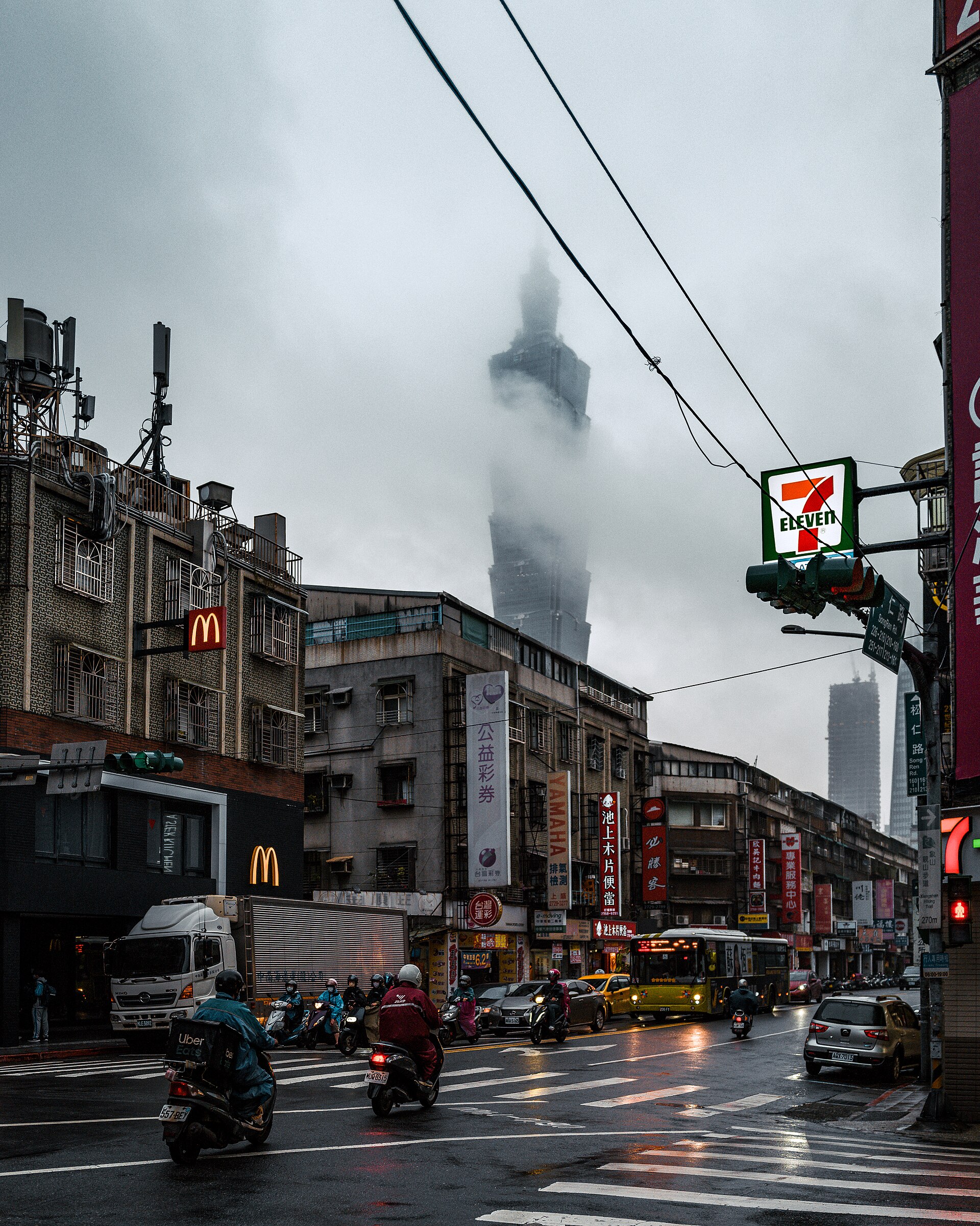 Low gray clouds partly obscure Taipei 101 above Songren Road, where scooter riders in rain gear travel through wet traffic.