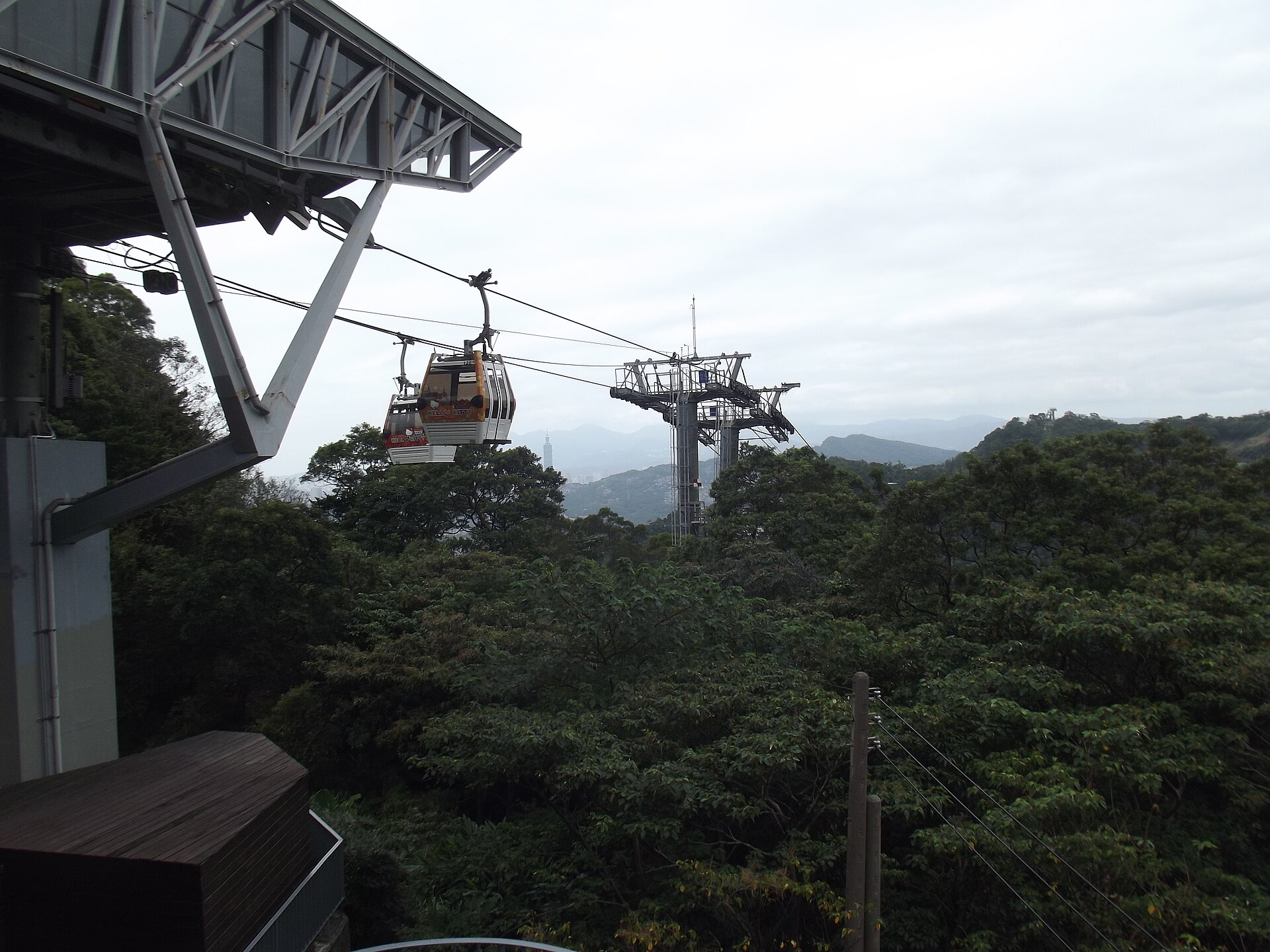 A Maokong Gondola cabin passes station structures above forested Taipei hills, with hazy city buildings in the distance.
