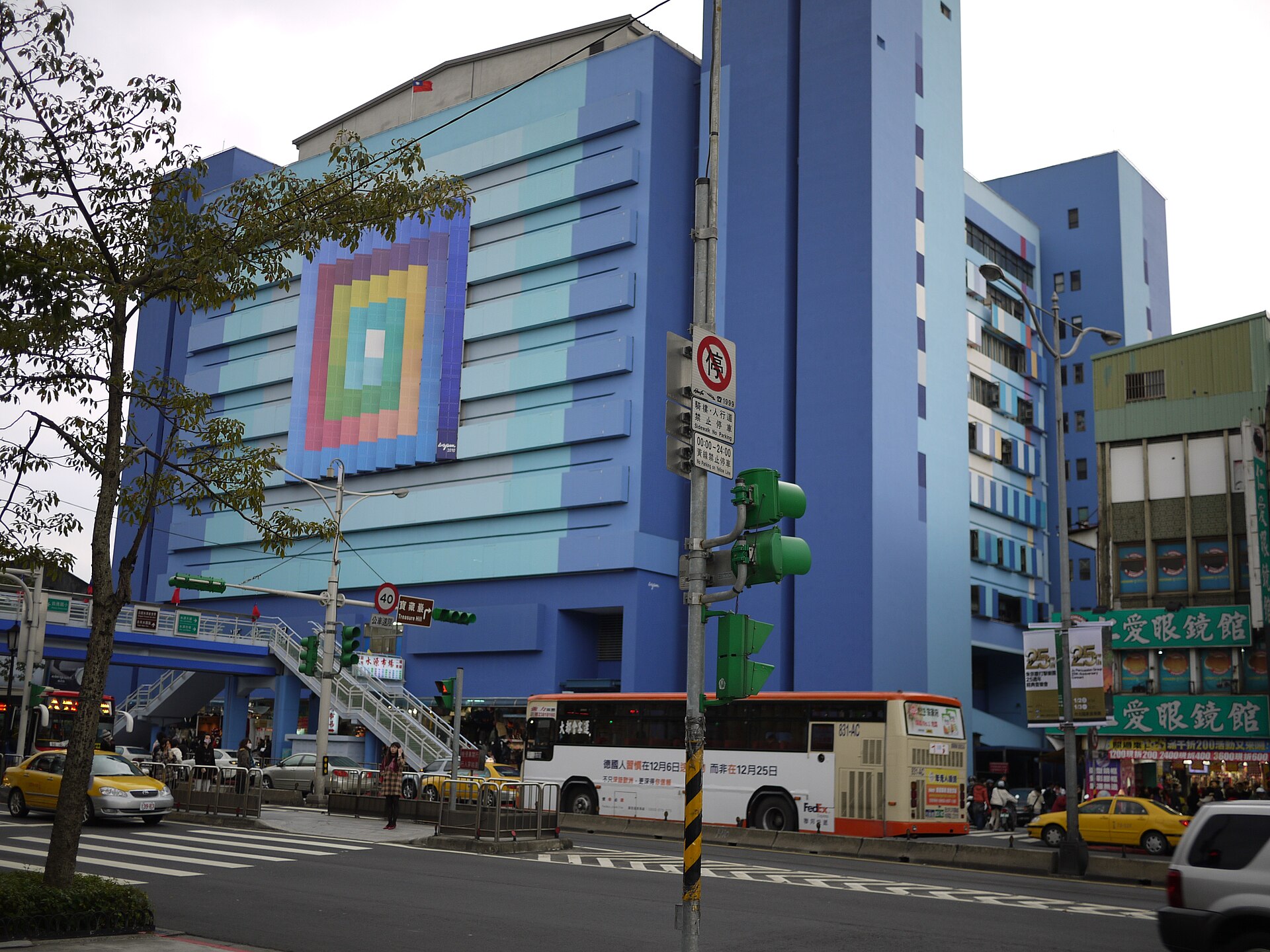 An orange and white city bus passes the colorful Shuiyuan Building in Taipei, alongside yellow taxis and pedestrians.