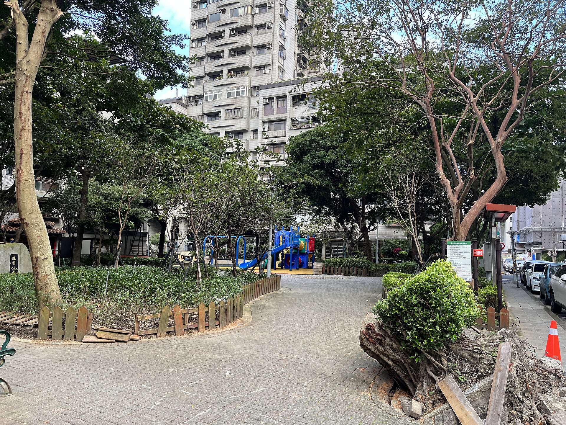 A path through trees and shrubs leads to blue playground equipment in a Taipei neighborhood park beside apartment buildings.
