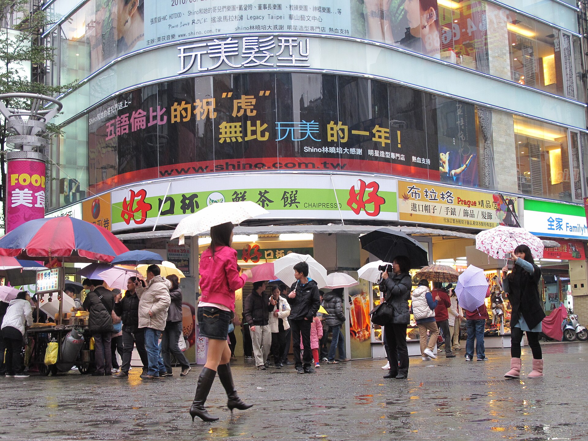 Pedestrians carry colorful umbrellas across wet paving in Taipei's Ximending shopping district, beneath large signs on a corner building.