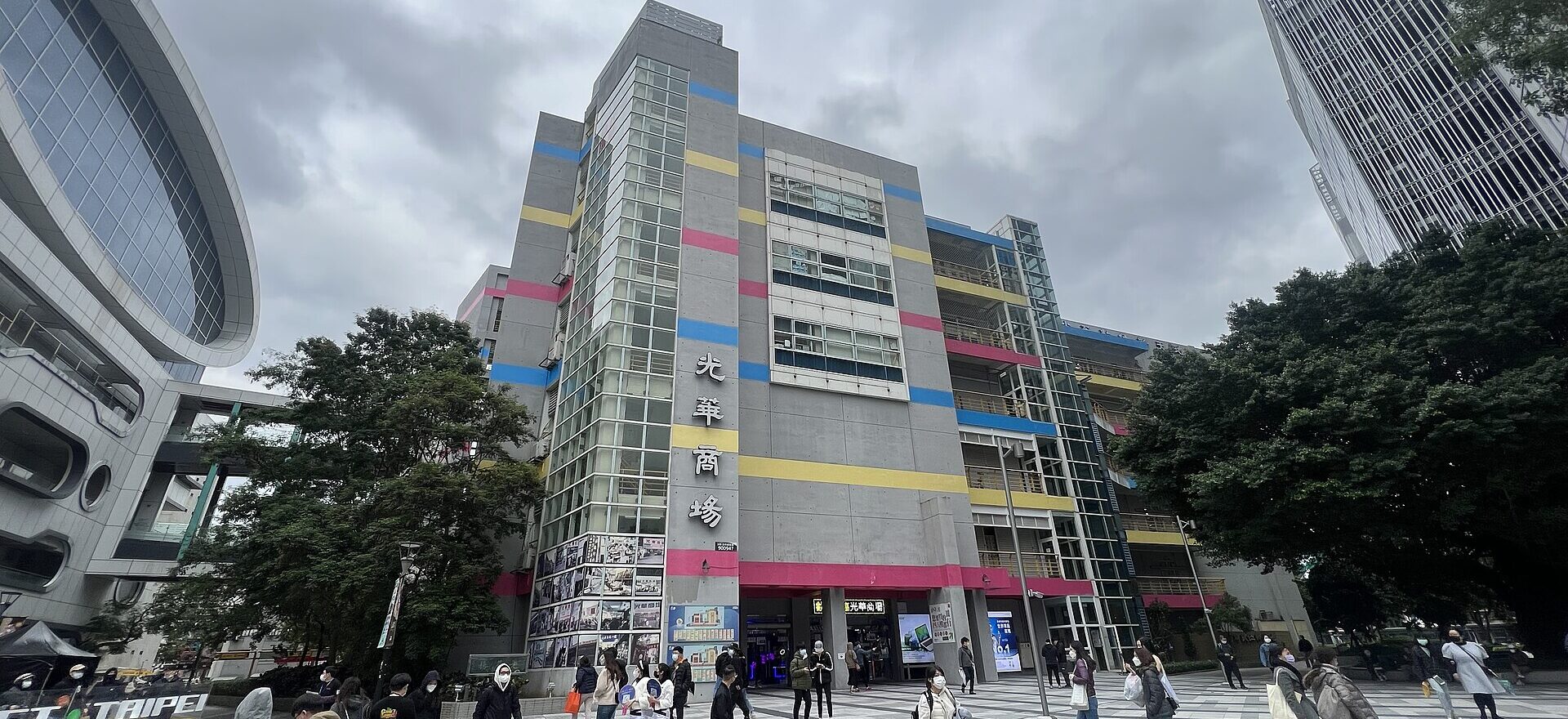 Pedestrians cross the plaza outside Guang Hua Digital Plaza in Taipei beneath its gray facade and colorful horizontal bands.