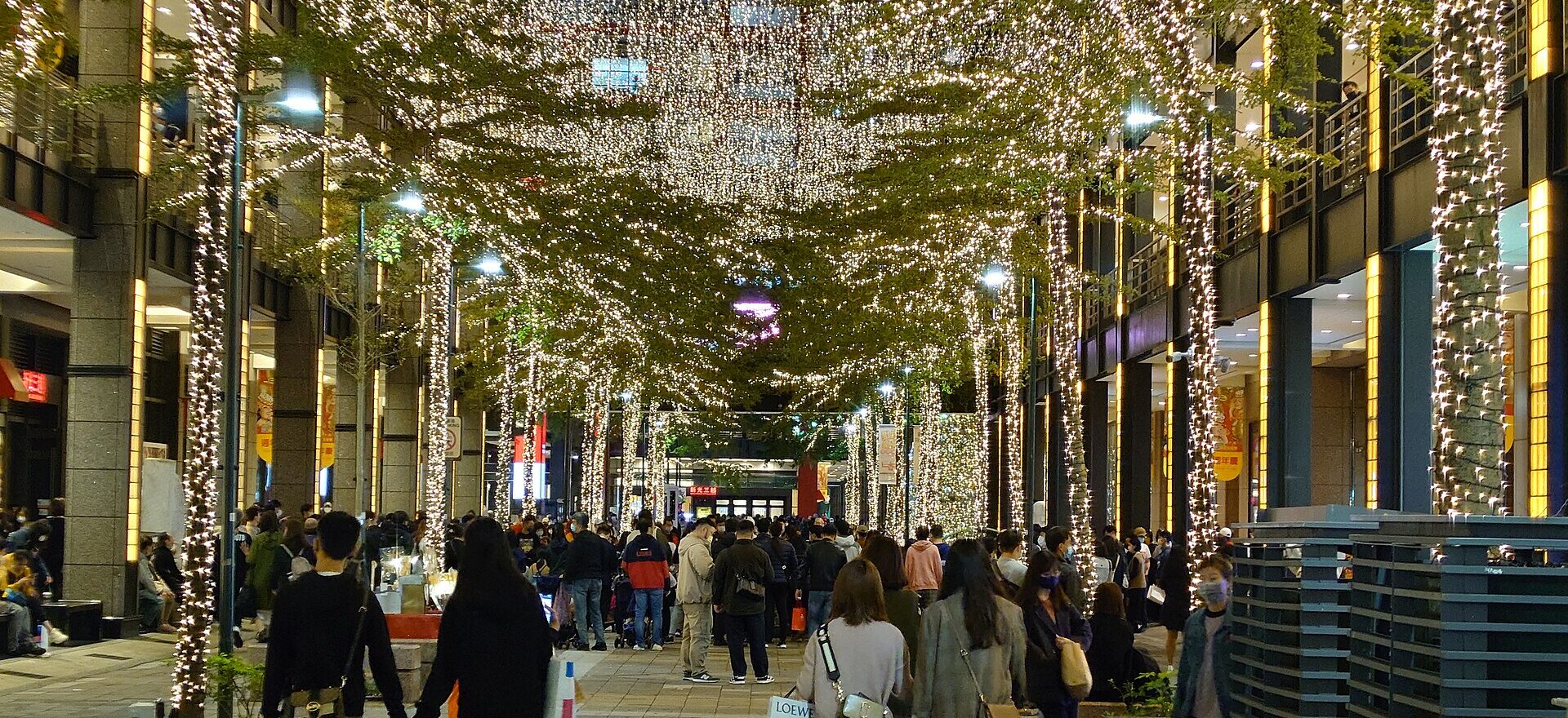 Pedestrians walk beneath trees wrapped in white lights at Chianti Plaza in Taipei, with illuminated shopping buildings on both sides.