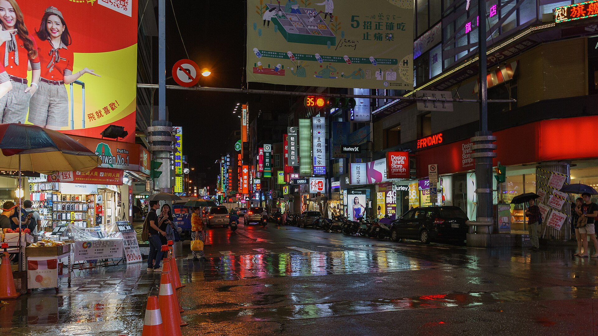 People shelter under umbrellas beside shops in Taipei's Ximending district, with colorful signs reflected across the wet street.