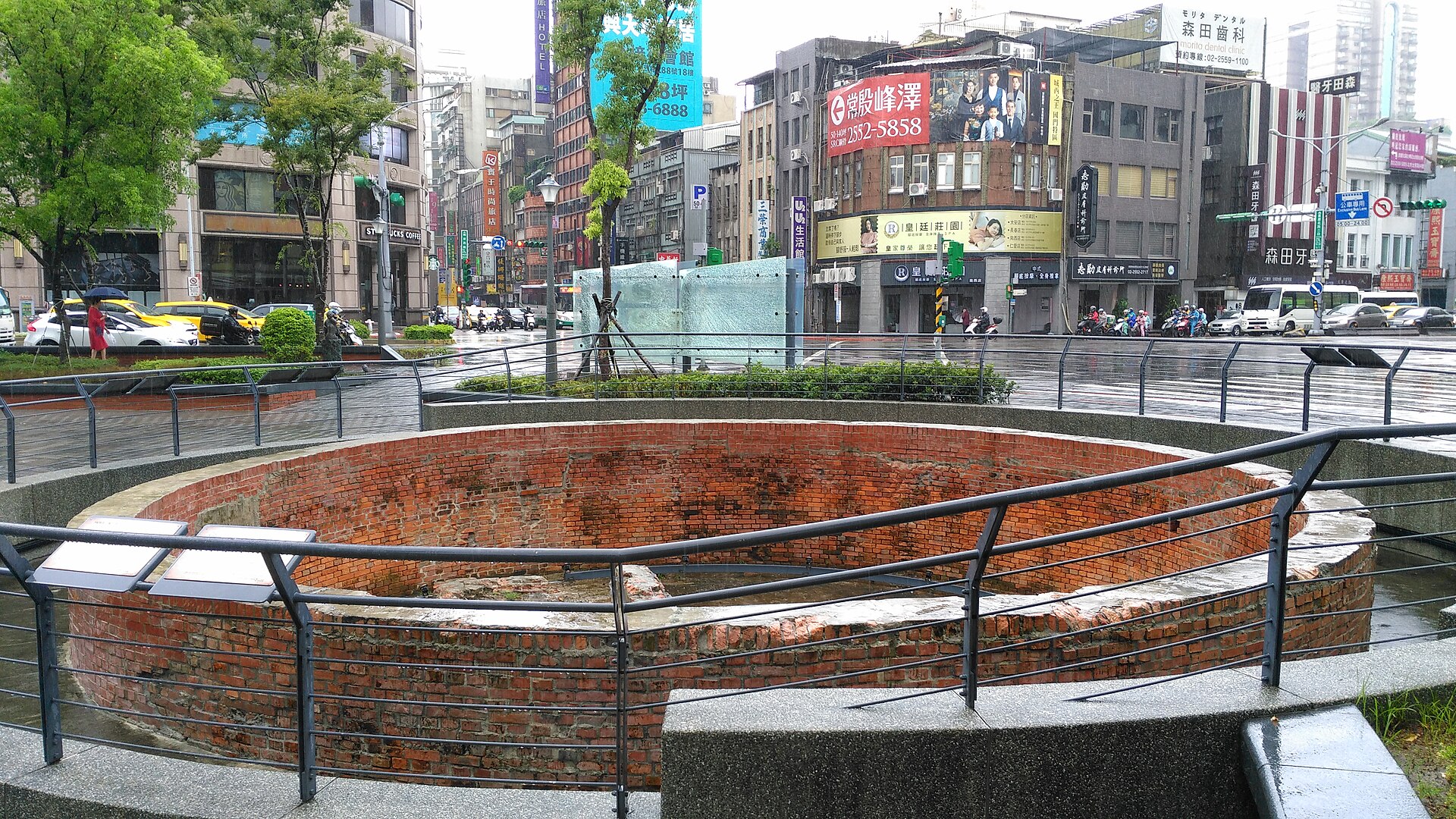 Rain falls over Dadaocheng Circle's brick cistern in Taipei, with wet streets, shops, taxis and scooter riders behind it.