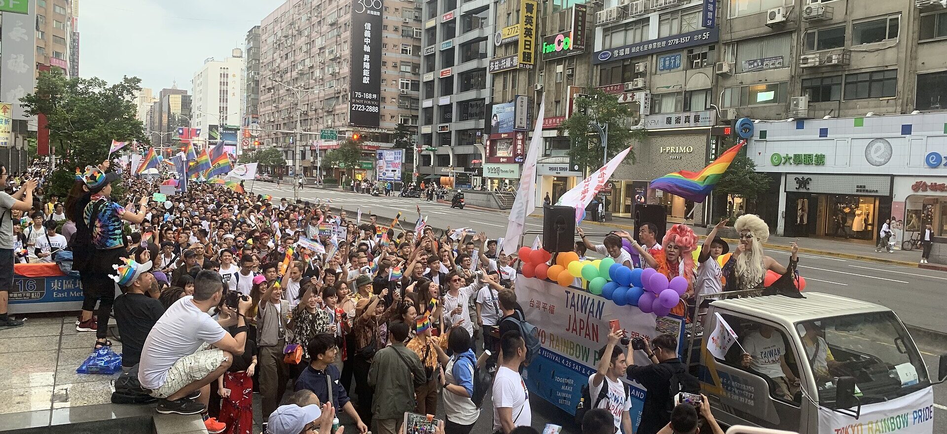 Rainbow flags and balloons surround a parade truck as a large Taipei Pride crowd fills the street between multistory buildings.
