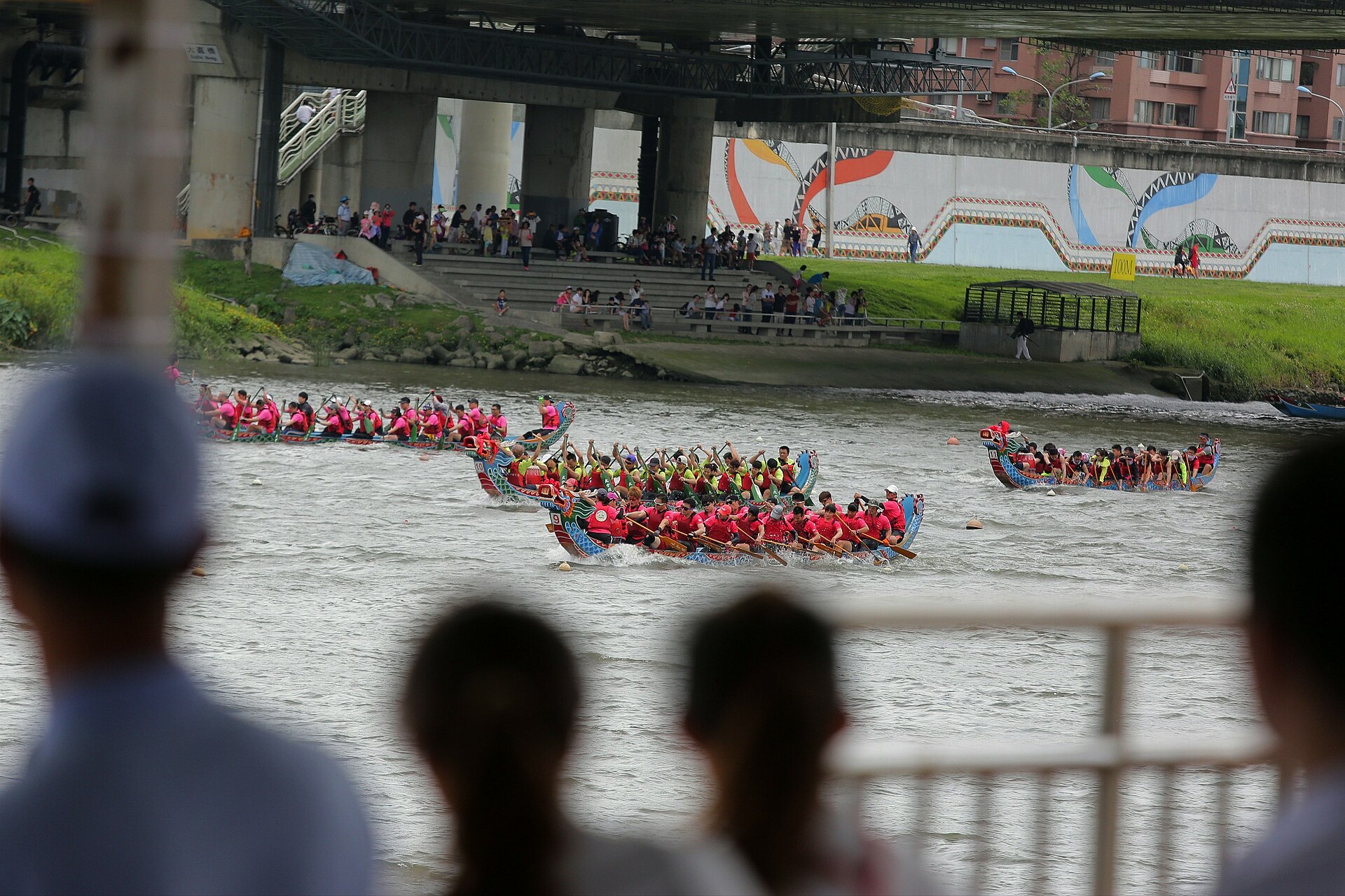 Several decorated dragon boats race along the Keelung River in Taipei, with spectators watching from both riverbanks.