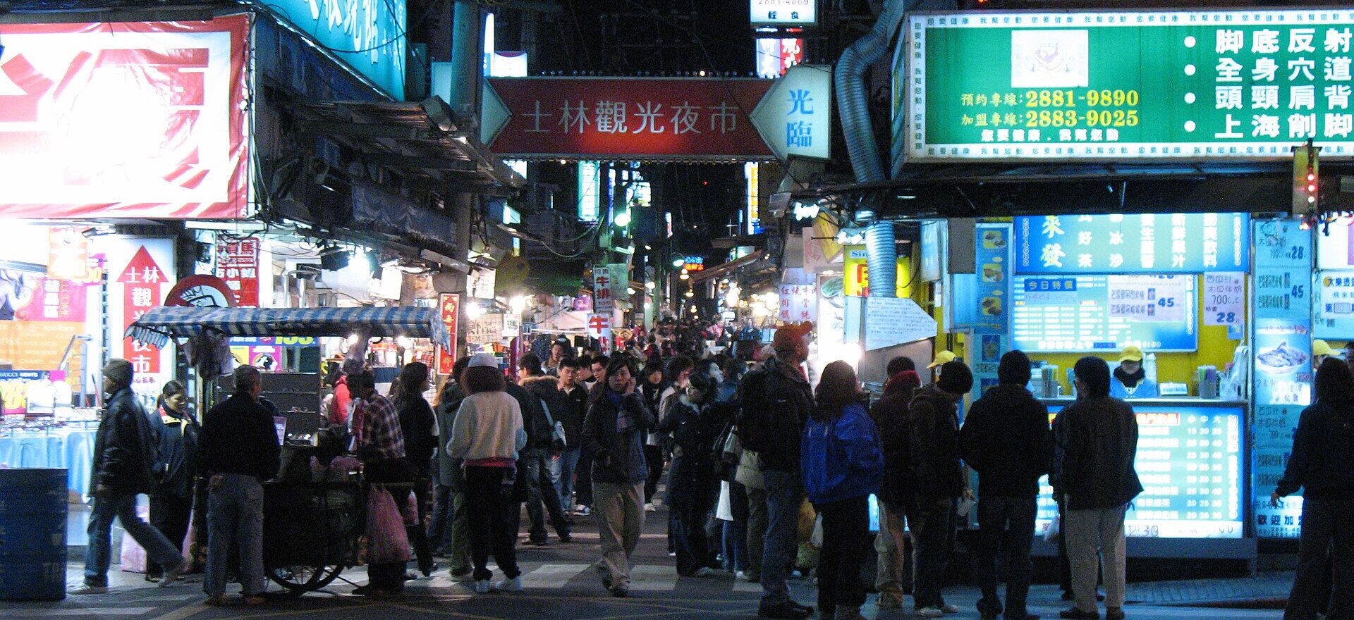 Shoppers crowd the brightly lit entrance to a Shilin Night Market lane in Taipei beneath an overhead market sign.