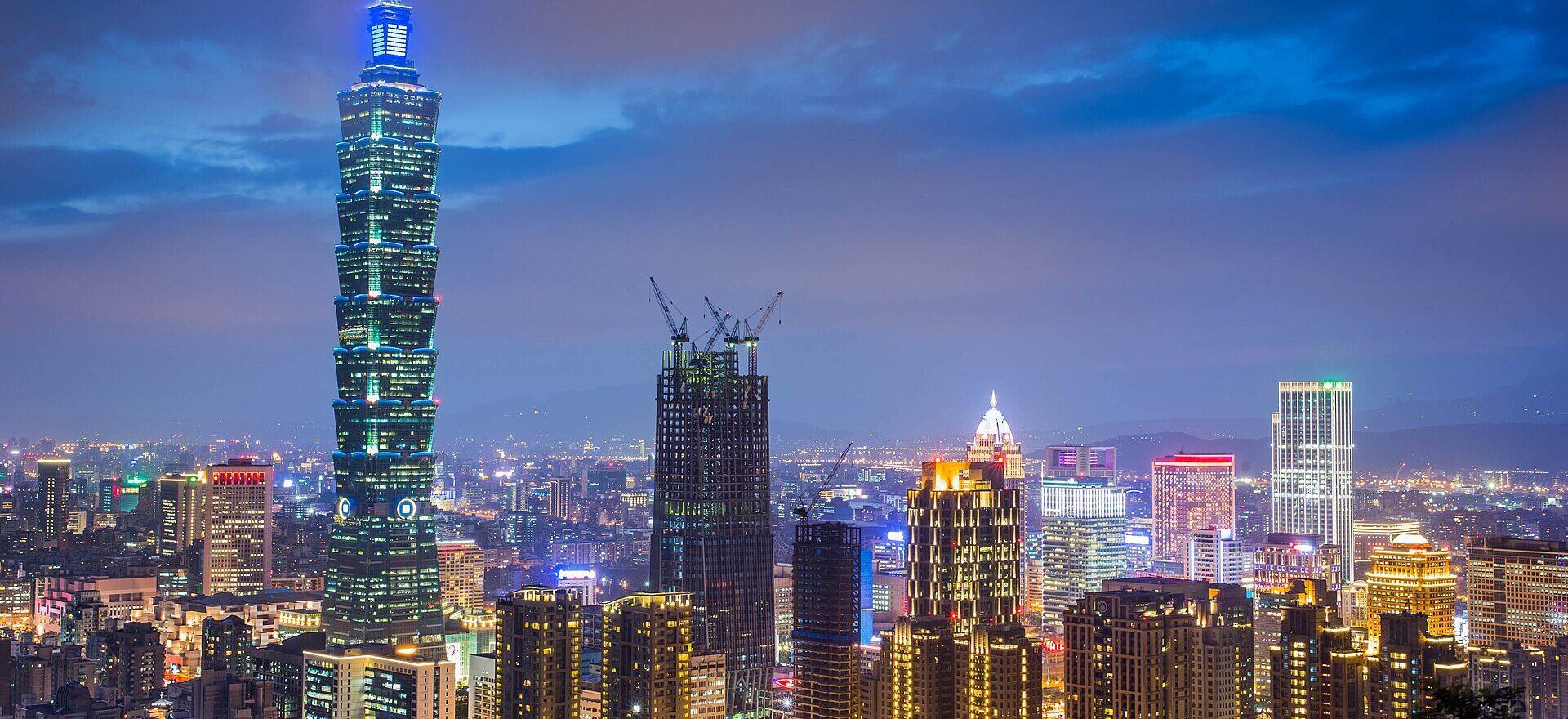 Taipei 101 rises above illuminated towers and streets in Xinyi District, Taipei, under a deep blue evening sky.