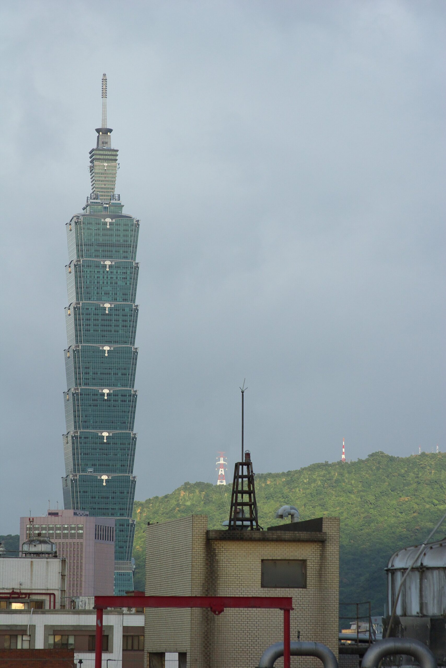 Taipei 101 towers above rooftop equipment and a green hillside beneath gray storm clouds in an August view of Taipei.