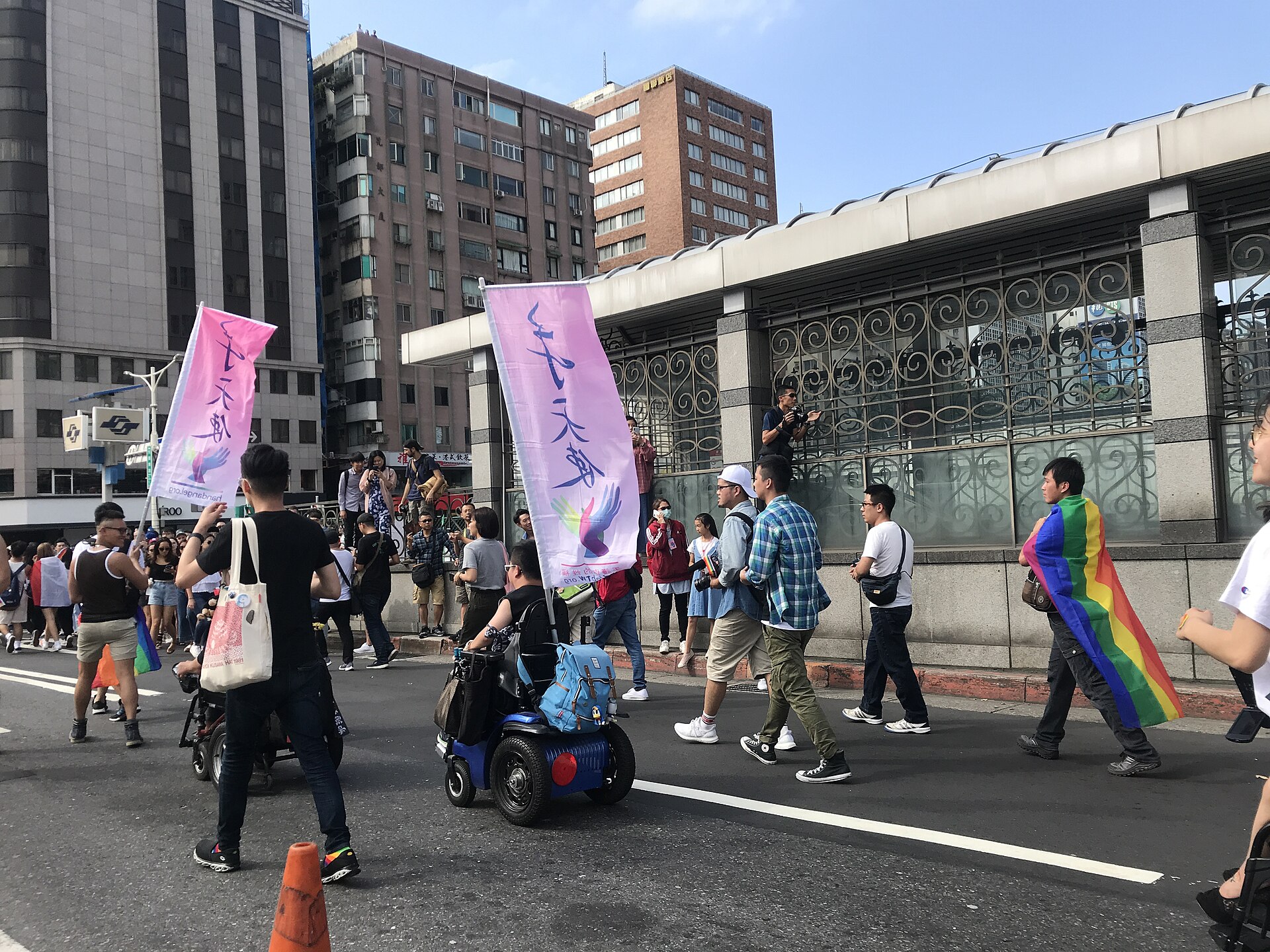 Taipei Pride participants march beside a metro entrance, including a mobility scooter user and a person draped in a rainbow flag.