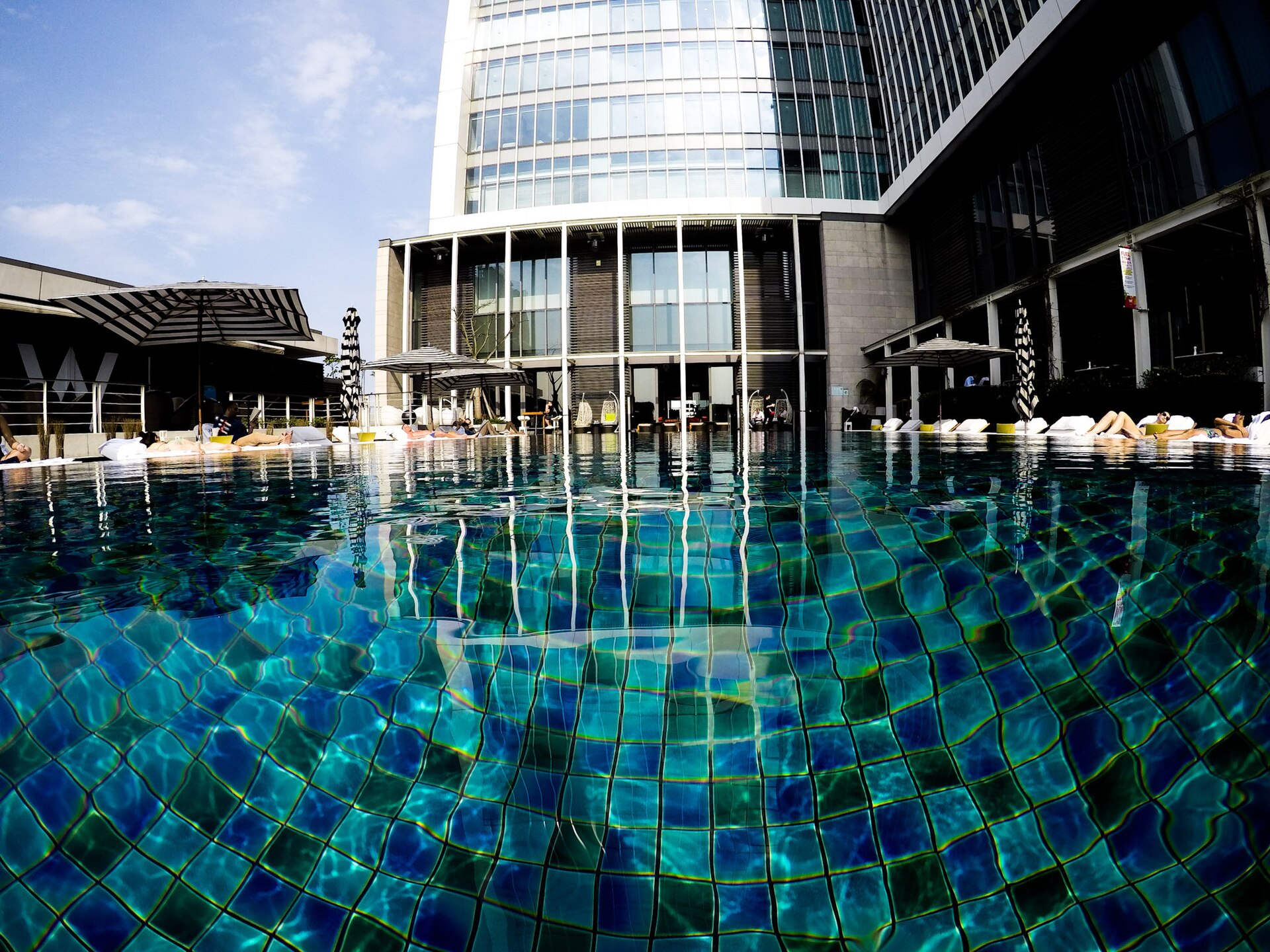 Tiled outdoor pool at W Taipei with striped parasols, sunbathers on loungers and the hotel's glass tower rising behind.
